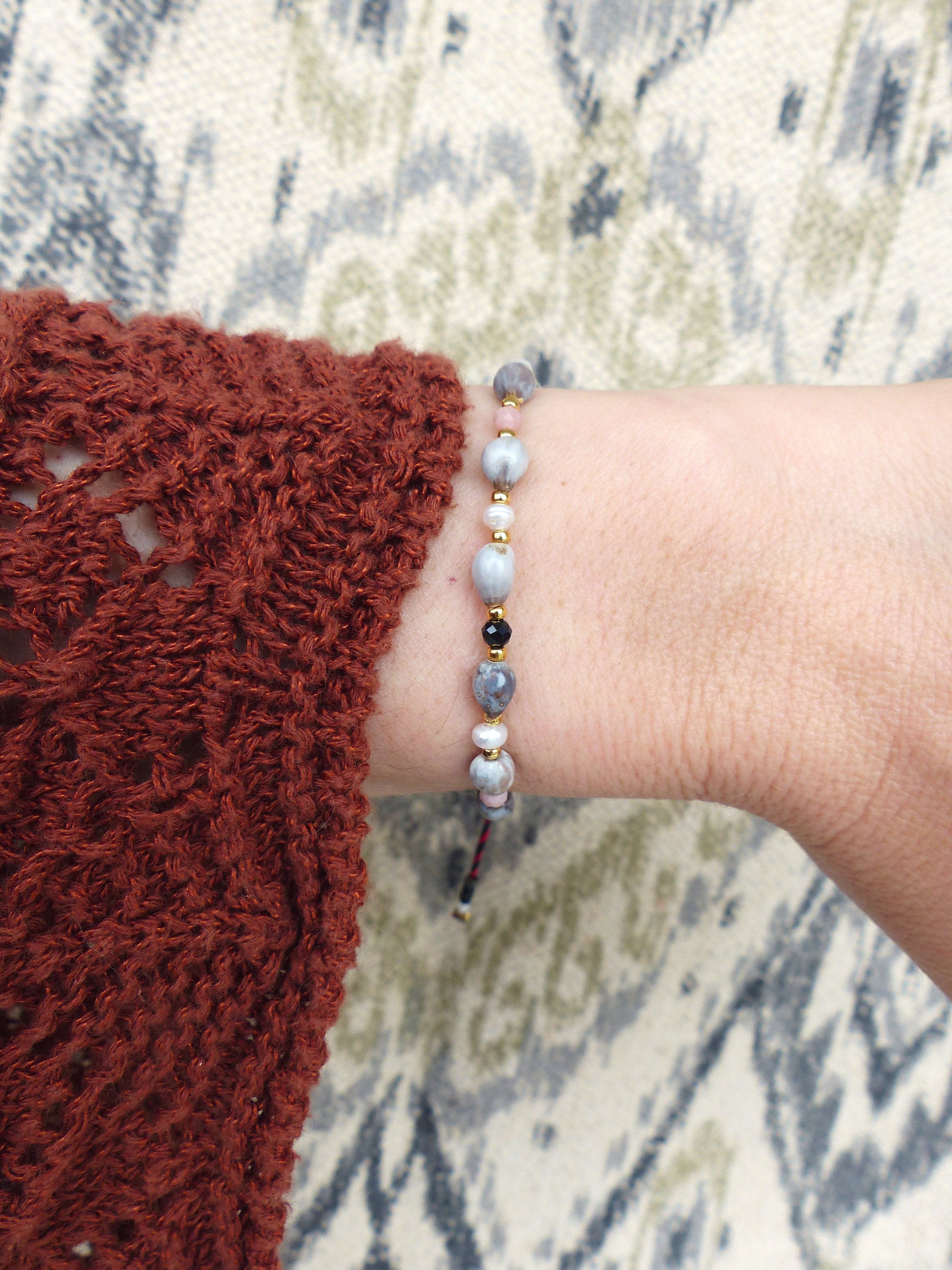 Peut inclure: Un bracelet avec des perles grises, noires et dorées sur une ficelle. Le bracelet est noué avec une ficelle rouge et blanche.