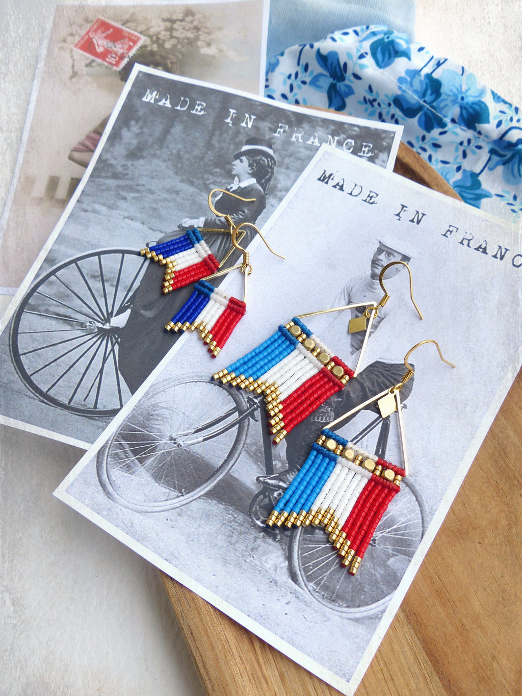 Peut inclure: Une paire de boucles d'oreilles dorées avec des franges de perles bleues, blanches et rouges, ressemblant au drapeau français. Les boucles d'oreilles sont présentées sur une carte postale vintage avec le texte "Made in France".