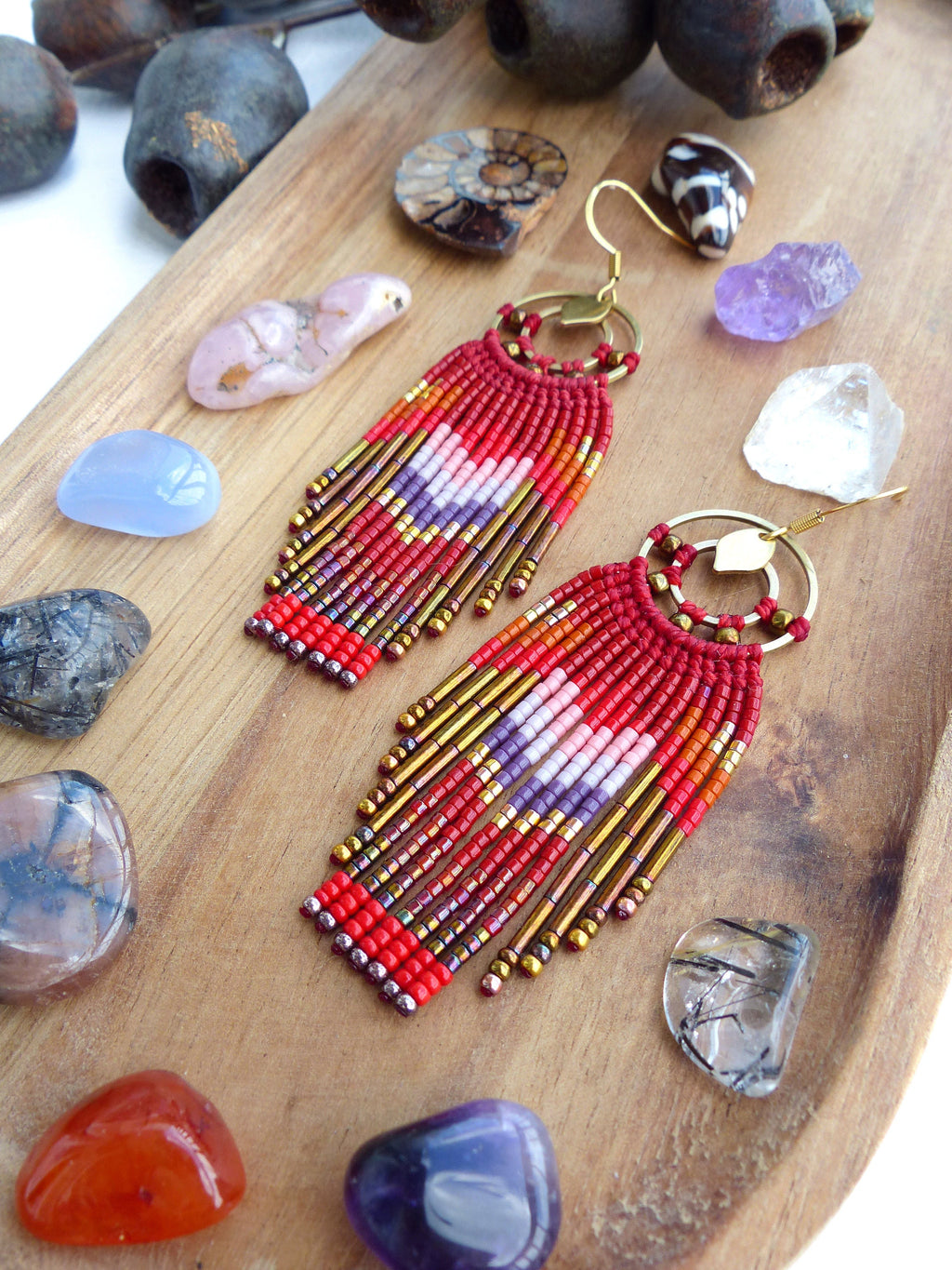 Peut inclure: Boucles d'oreilles en franges de perles rouges, blanches et dorées avec un design de cerceau doré. Les boucles d'oreilles sont présentées sur une surface en bois avec diverses pierres.