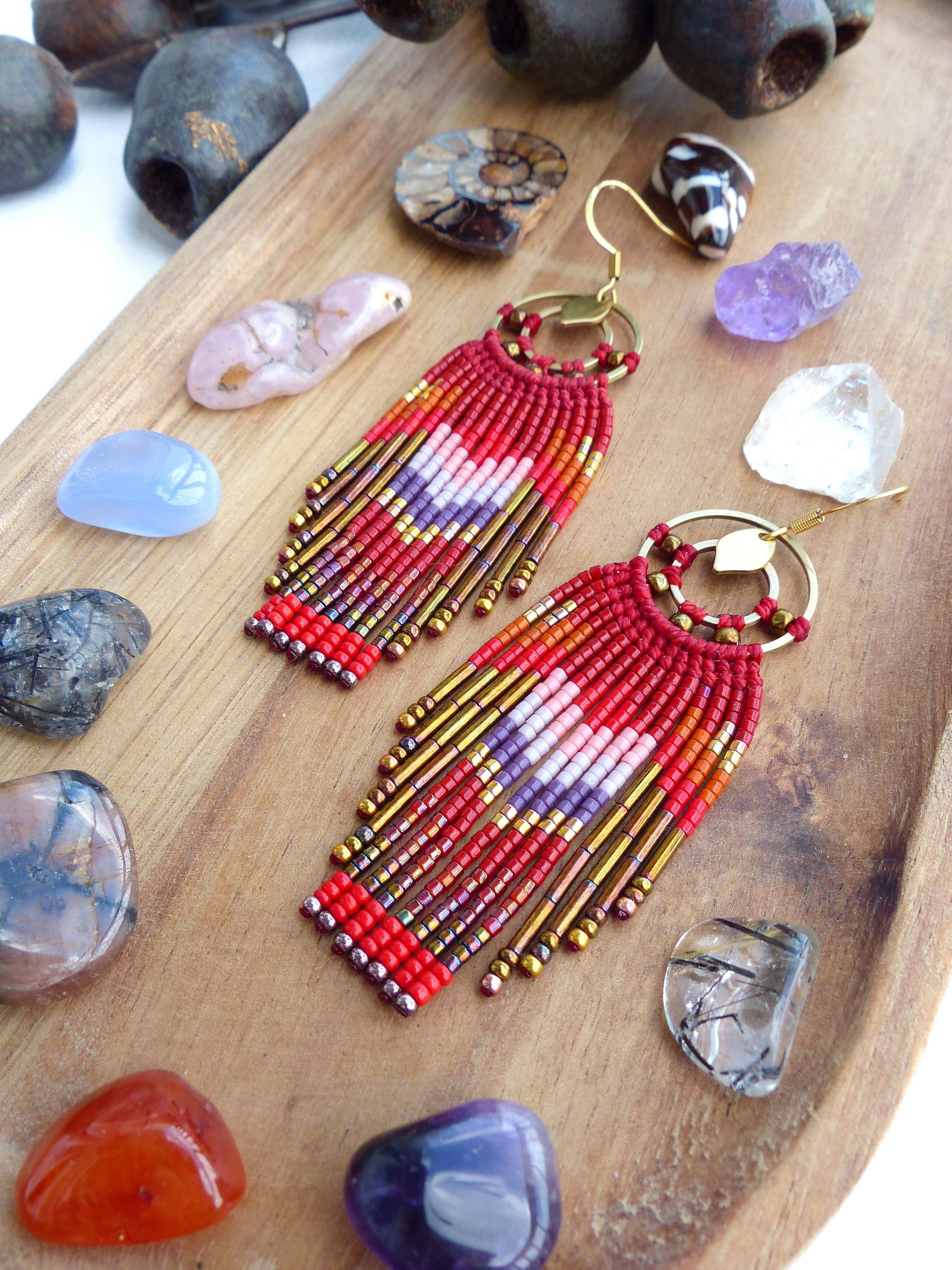 Peut inclure: Boucles d'oreilles en franges de perles rouges, blanches et dorées avec un design de cerceau doré. Les boucles d'oreilles sont présentées sur une surface en bois avec diverses pierres.