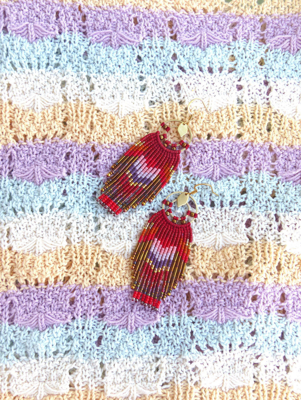 Peut inclure: Une paire de boucles d'oreilles pendantes en perles rouges, violettes et dorées avec des crochets dorés. Les boucles d'oreilles sont photographiées sur un tissu tricoté multicolore.