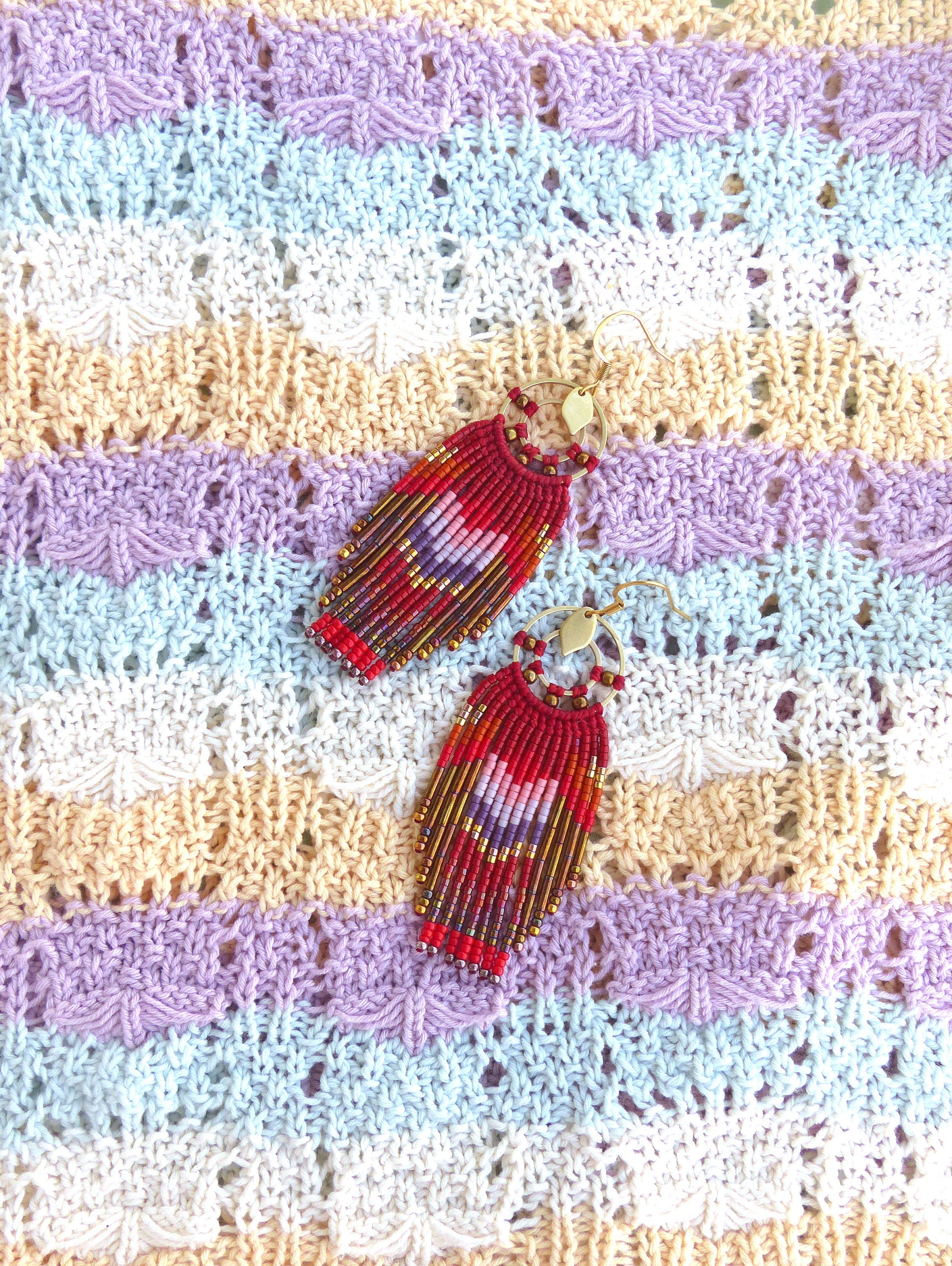 Peut inclure: Une paire de boucles d'oreilles pendantes en perles rouges, violettes et dorées avec des crochets dorés. Les boucles d'oreilles sont photographiées sur un tissu tricoté multicolore.