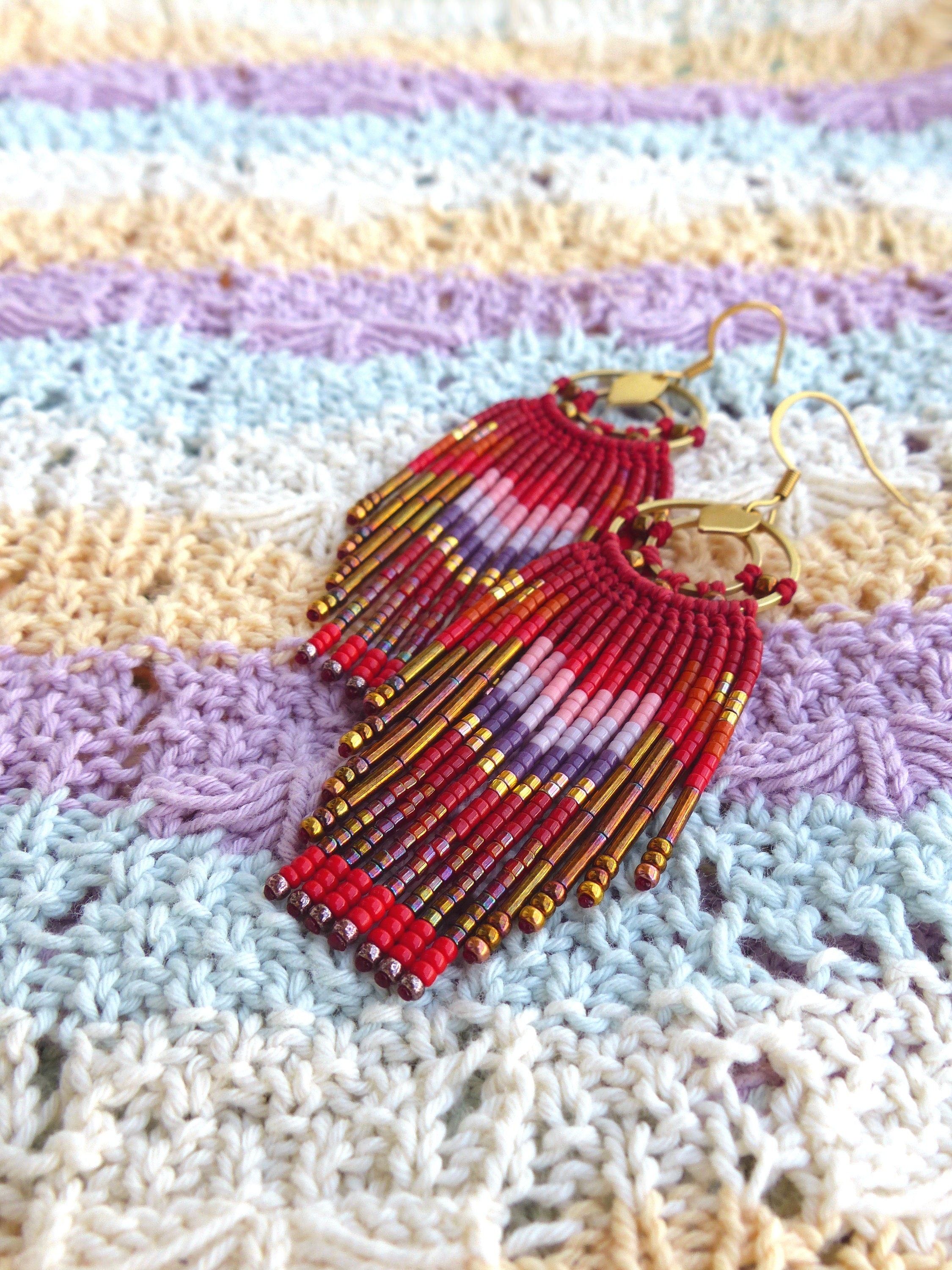 Peut inclure: Une paire de boucles d'oreilles en perles rouges, dorées et violettes avec un design en frange. Les boucles d'oreilles sont suspendues à un tissu crocheté rayé.