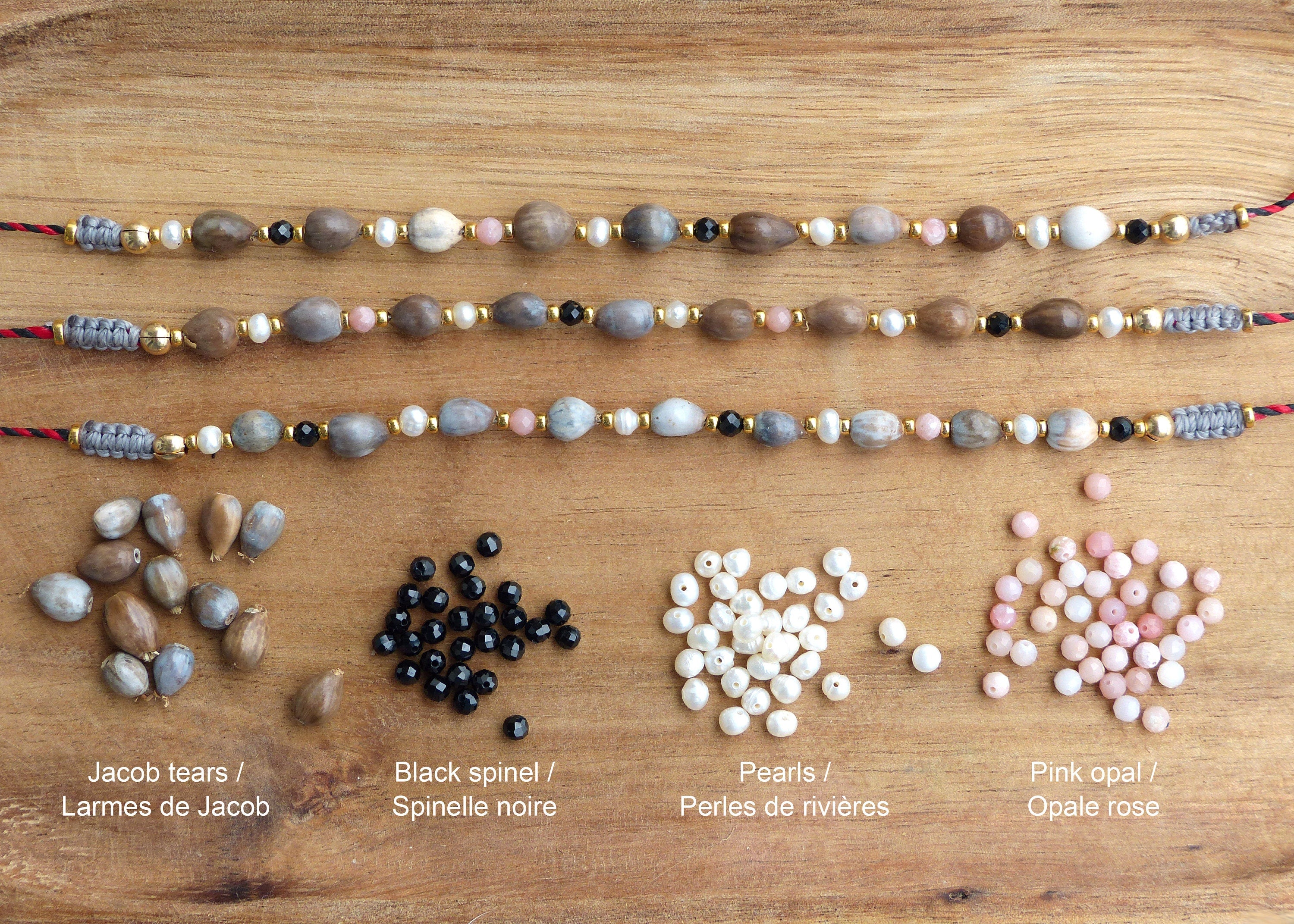 Peut inclure: Trois rangs de bijoux avec différentes perles, dont des perles brunes, grises, roses, blanches et noires. Les perles sont disposées en rangée sur une surface en bois. En dessous des bijoux se trouvent des tas de perles en vrac des mêmes couleurs. Le texte sous les perles indique "Jacob tears / Larmes de Jacob", "Black spinel / Spinelle noire", "Pearls / Perles de rivières", et "Pink opal / Opale rose".