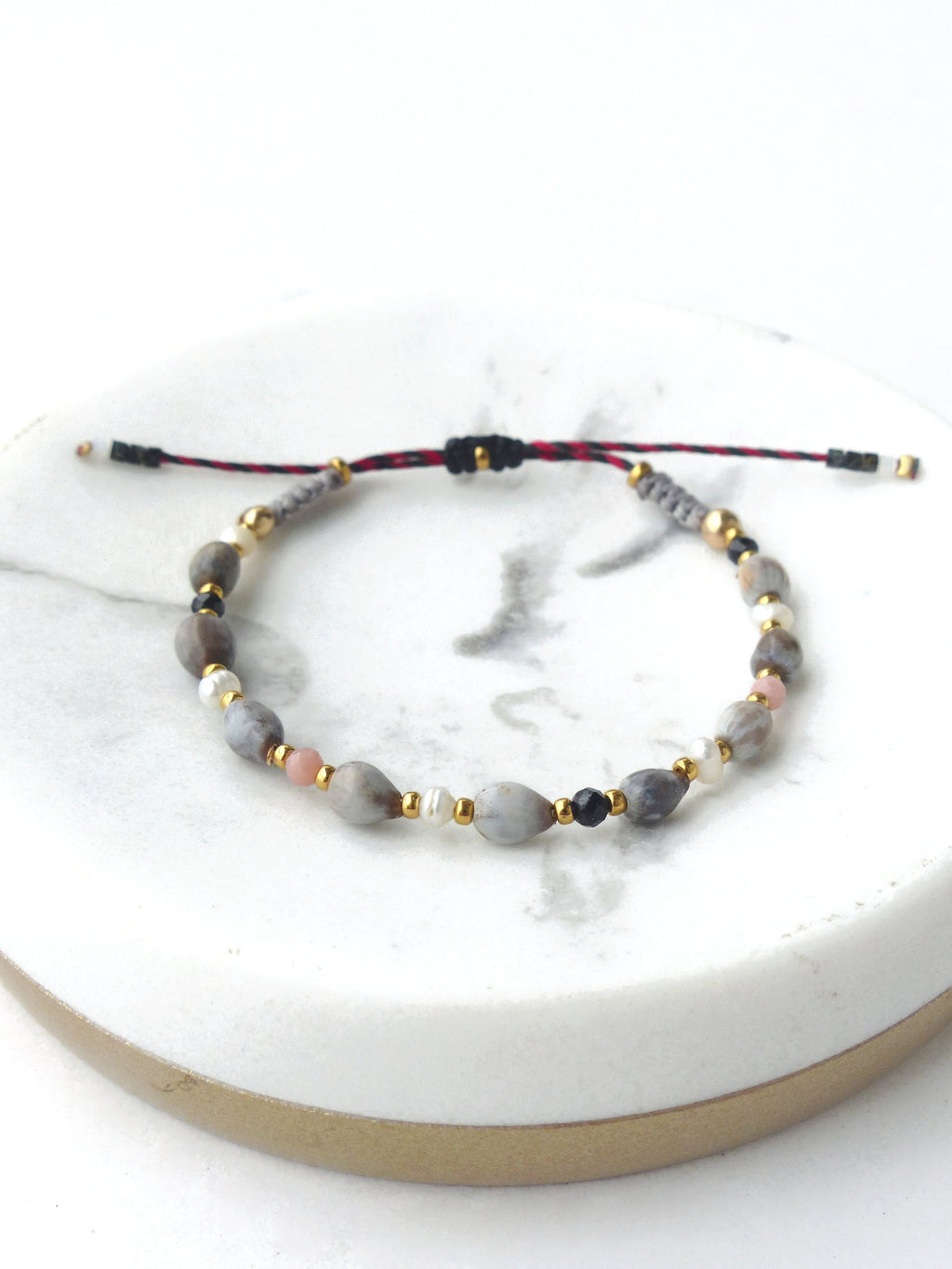 Peut inclure: Un bracelet composé de perles de différentes couleurs, notamment grises, blanches, roses et noires. Le bracelet est réalisé avec un cordon rouge et noir et possède un nœud coulissant.