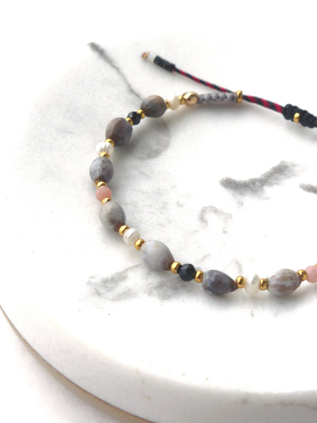 Peut inclure: Un bracelet composé de perles grises, blanches, roses et noires. Les perles sont enfilées sur un cordon noir et rouge avec des accents dorés.