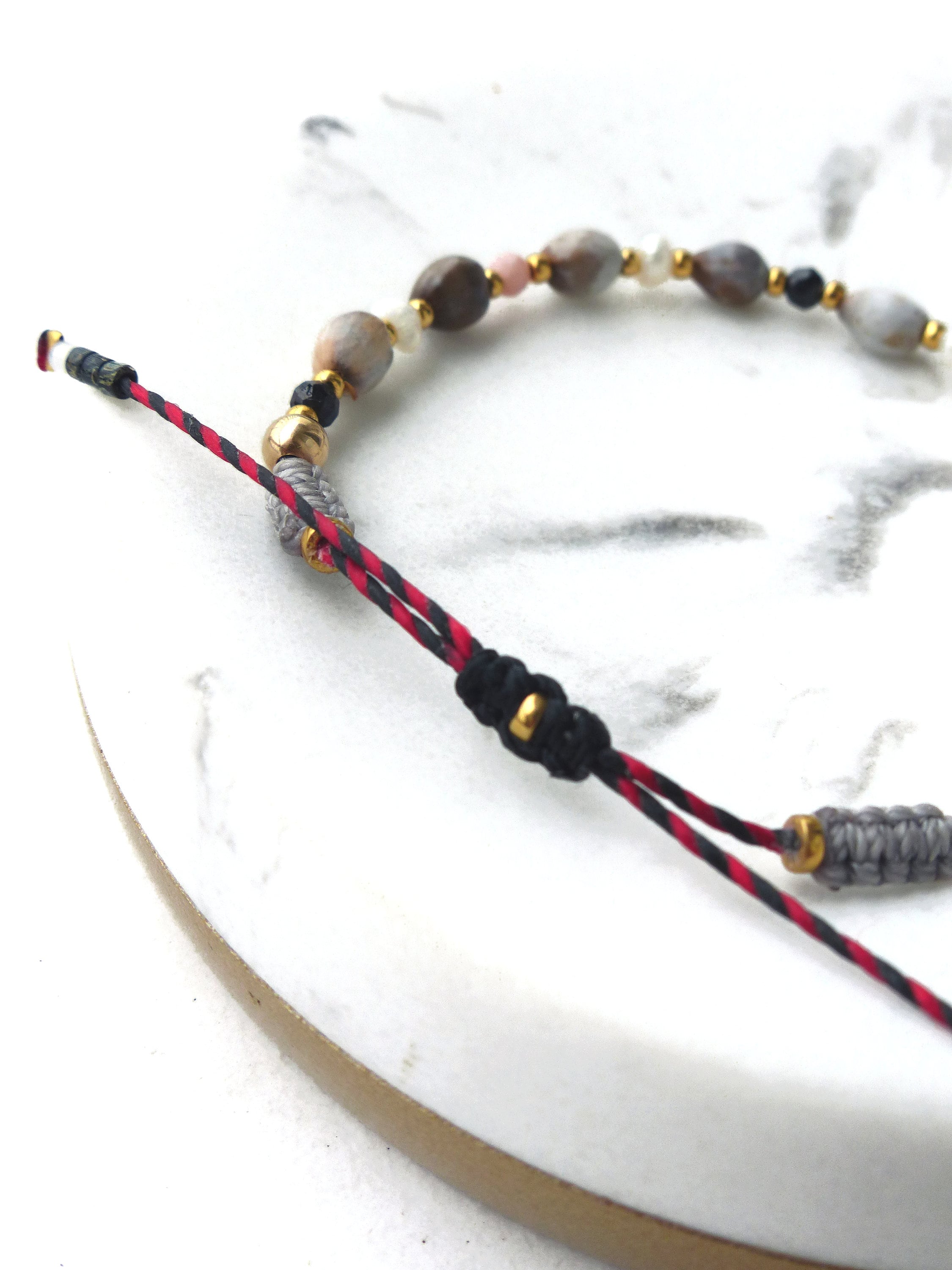 Peut inclure: Un bracelet avec un cordon rouge et noir, des perles grises et dorées, et une variété de perles de pierre naturelle. Le bracelet est noué et a une petite perle dorée à l'extrémité.