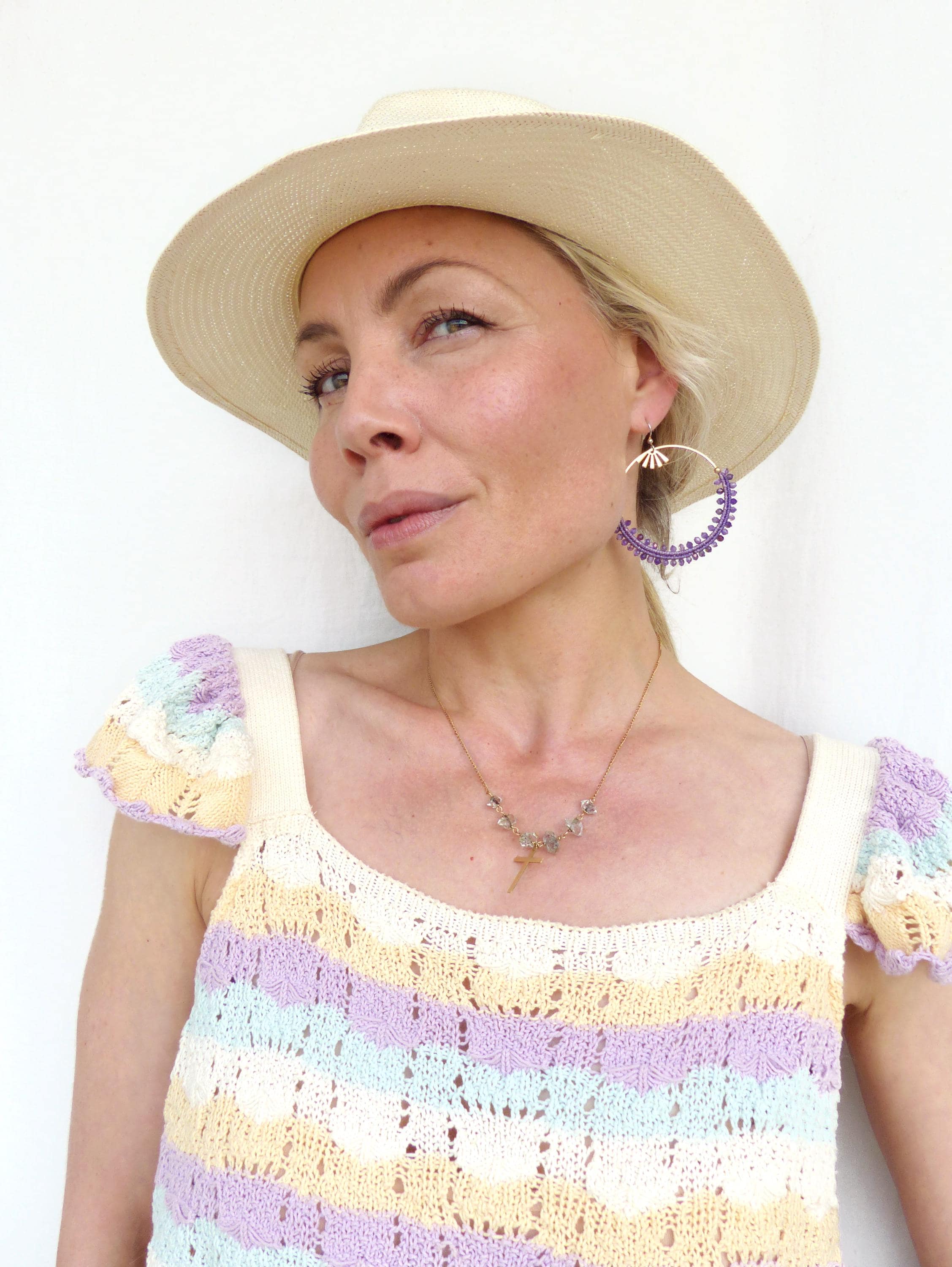 Peut inclure: Une femme porte un chapeau crème, un haut en crochet à rayures pastel et de grandes boucles d'oreilles créoles violettes. Un collier en or avec un pendentif en forme de croix est également visible. Le haut a une encolure carrée et des manches courtes.