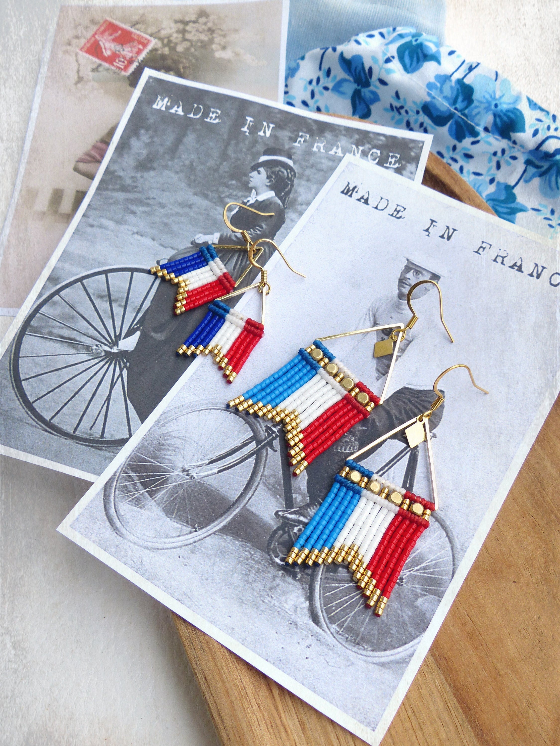 Peut inclure: Une paire de boucles d'oreilles dorées avec des franges de perles bleues, blanches et rouges, ressemblant au drapeau français. Les boucles d'oreilles sont présentées sur une carte postale vintage avec le texte "Made in France".