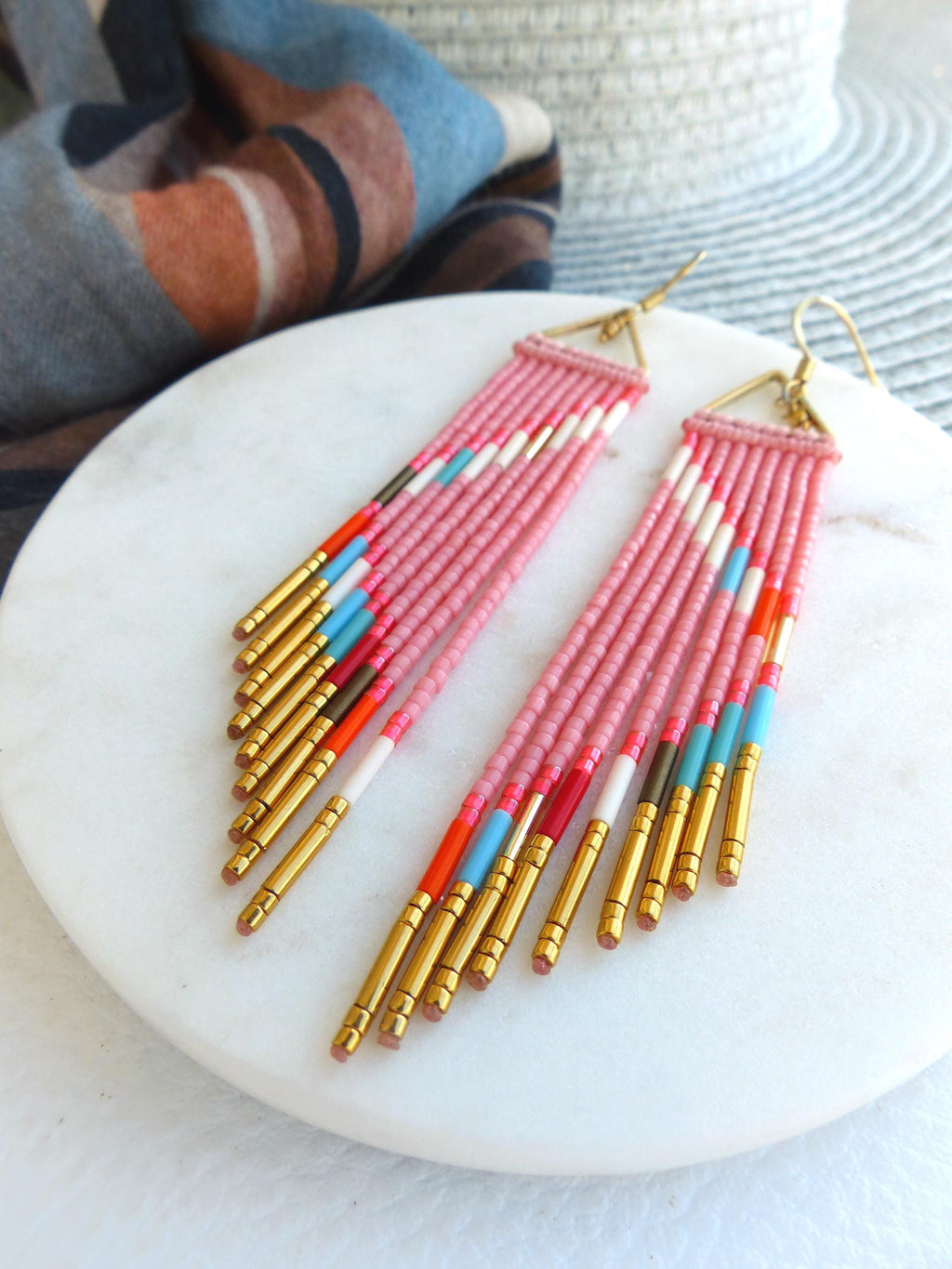 Peut inclure: Boucles d'oreilles pendantes roses en perles avec des accents dorés. Elles présentent un dessus triangulaire et plusieurs rangées de perles roses avec des rayures colorées blanches, rouges, oranges et bleues. Les extrémités sont dorées.