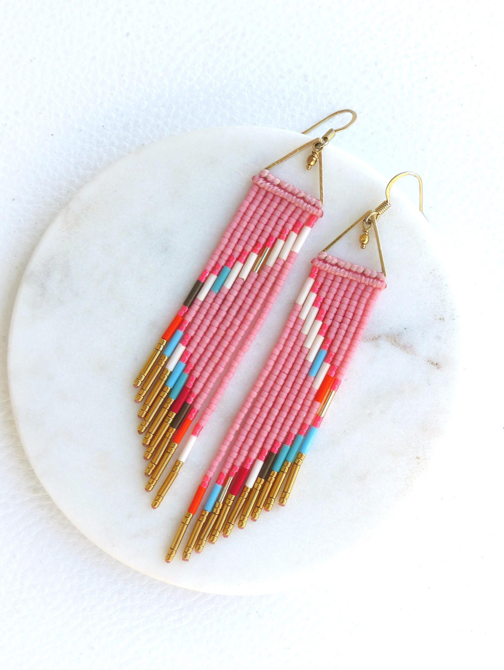 Peut inclure: Une paire de boucles d'oreilles roses en perles avec un motif géométrique. Les boucles d'oreilles ont un dessus triangulaire doré et des franges dorées. Le motif comprend des perles blanches, rouges, bleues et noires.
