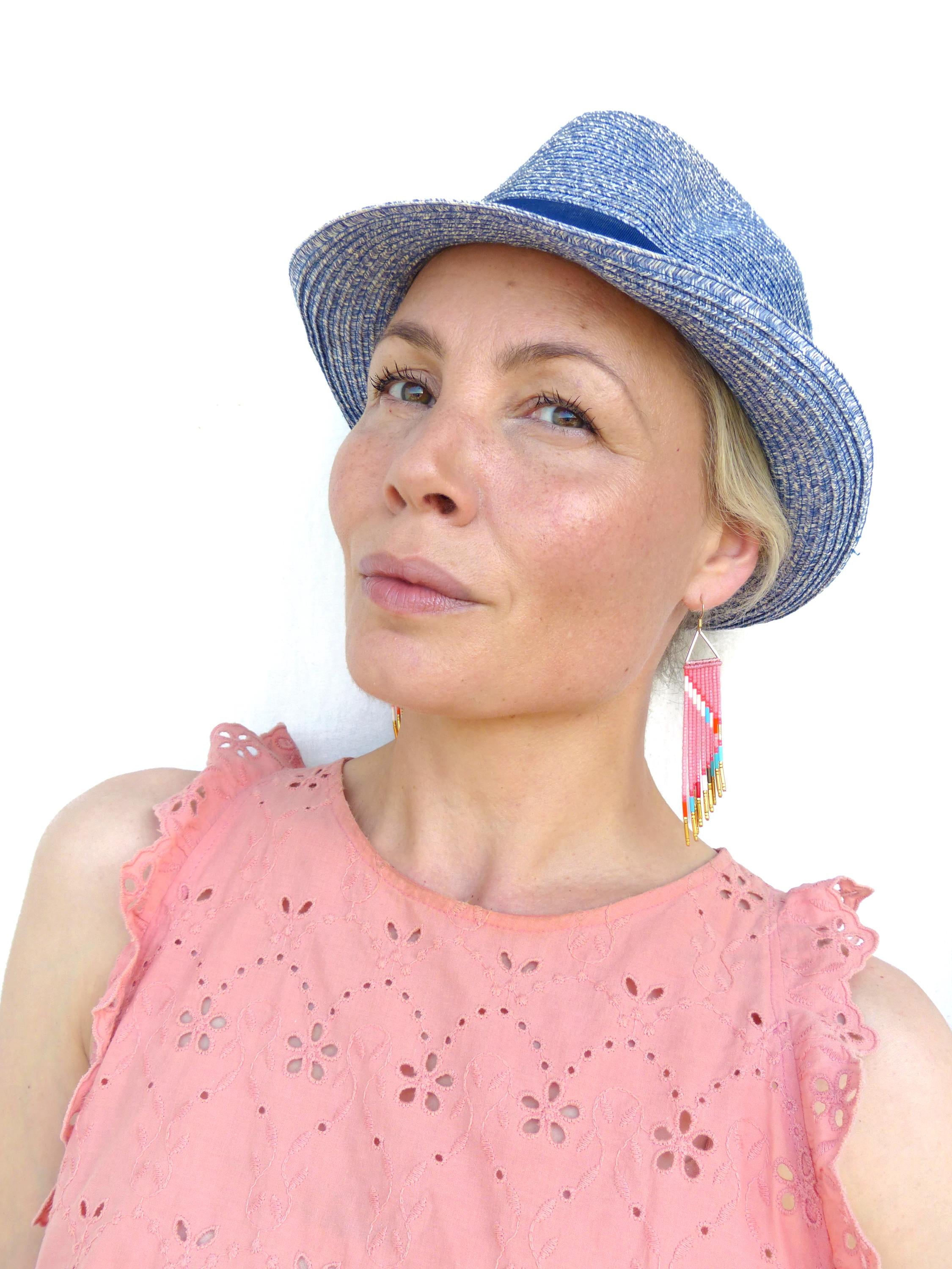 Peut inclure: Un chapeau fedora bleu clair avec un ruban bleu. Boucles d'oreilles roses en perles avec un motif géométrique et des accents dorés. Un haut rose à œillets avec des découpes florales. La femme regarde vers la caméra.