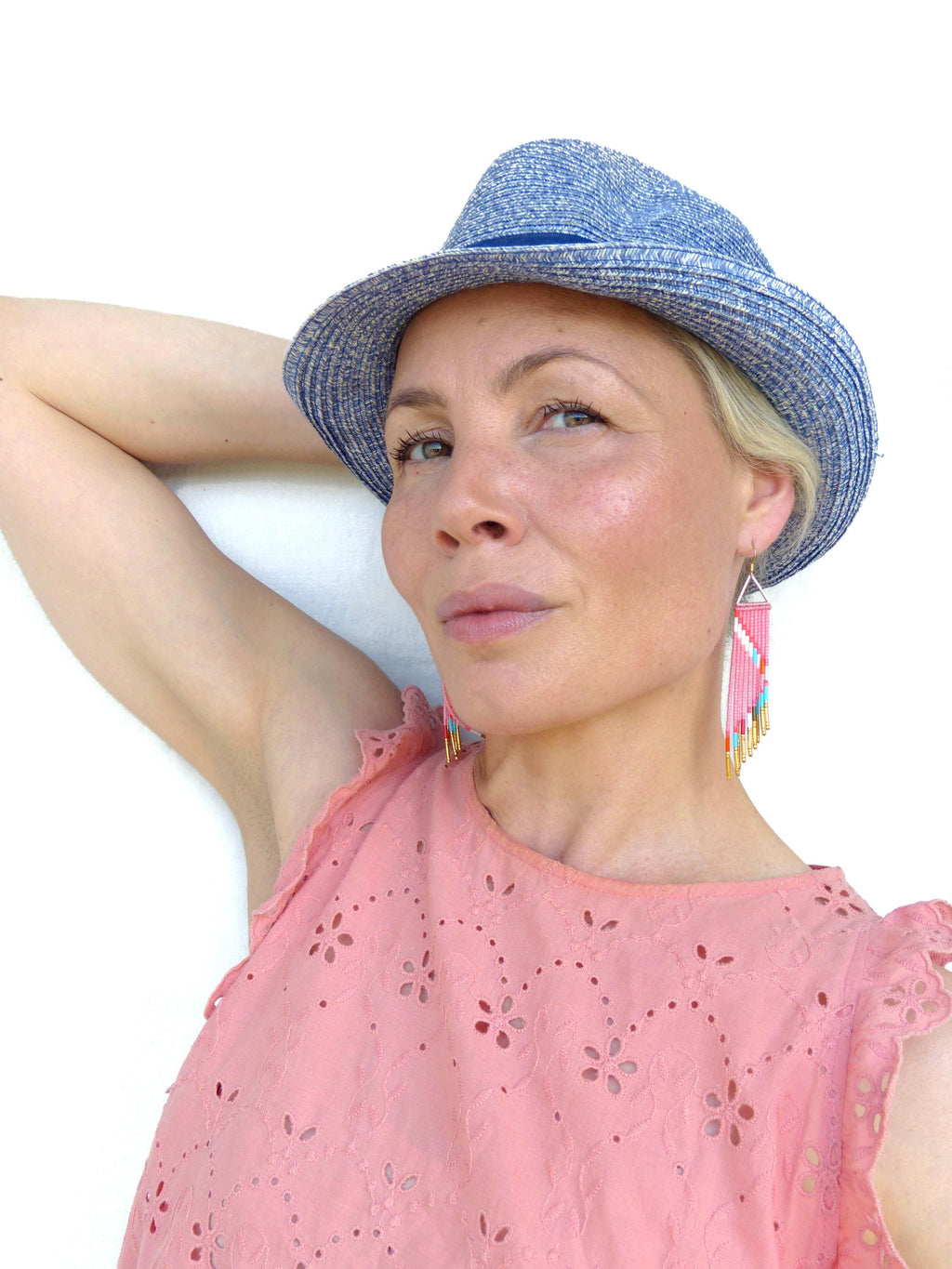Peut inclure: Une femme porte un chapeau de paille bleu et des boucles d'oreilles roses. Les boucles d'oreilles sont triangulaires avec un motif de perles roses et blanches et des franges dorées. Elle porte un haut rose avec des découpes florales. Le fond est blanc.