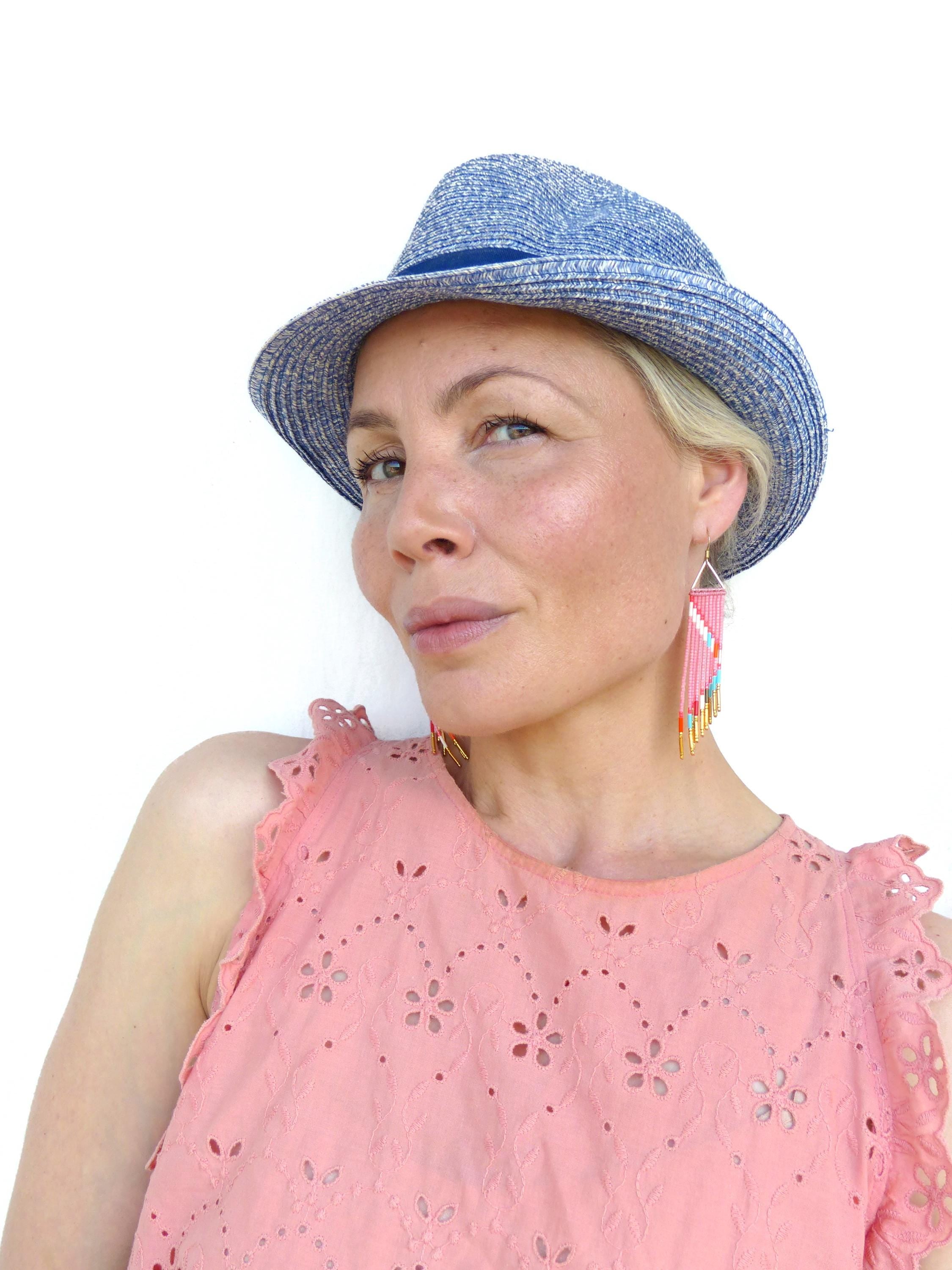 Peut inclure: Une femme porte un chapeau fedora bleu et des boucles d'oreilles roses. Les boucles d'oreilles sont triangulaires avec des perles roses, blanches et turquoise. Elle porte un haut sans manches rose avec un motif ajouré floral.