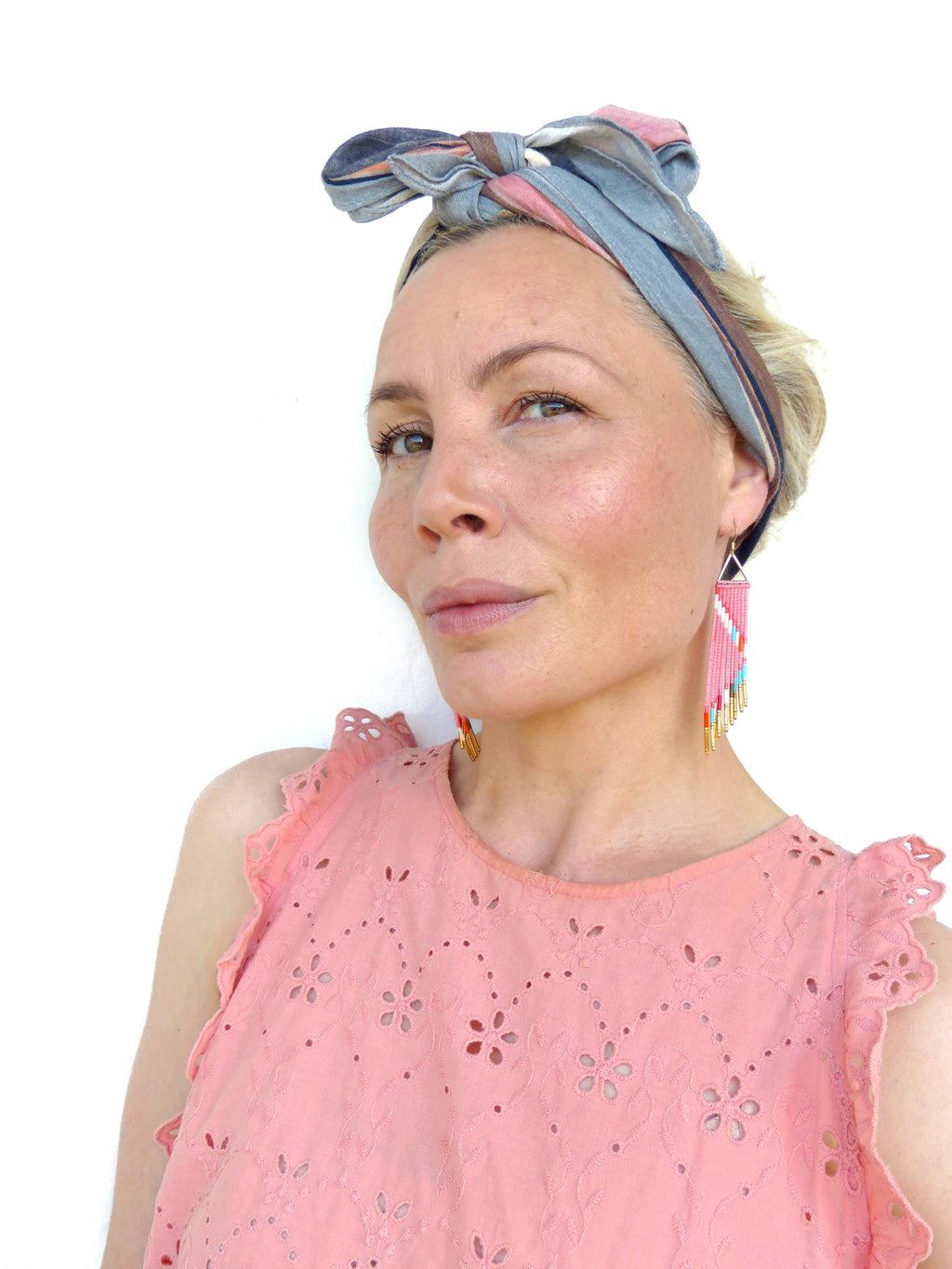 Peut inclure: Haut rose avec découpes florales et manches à volants. La personne porte un bandeau rayé et des boucles d'oreilles en perles avec des perles roses, blanches et turquoise. Les boucles d'oreilles ont un dessus triangulaire et une frange de perles colorées.