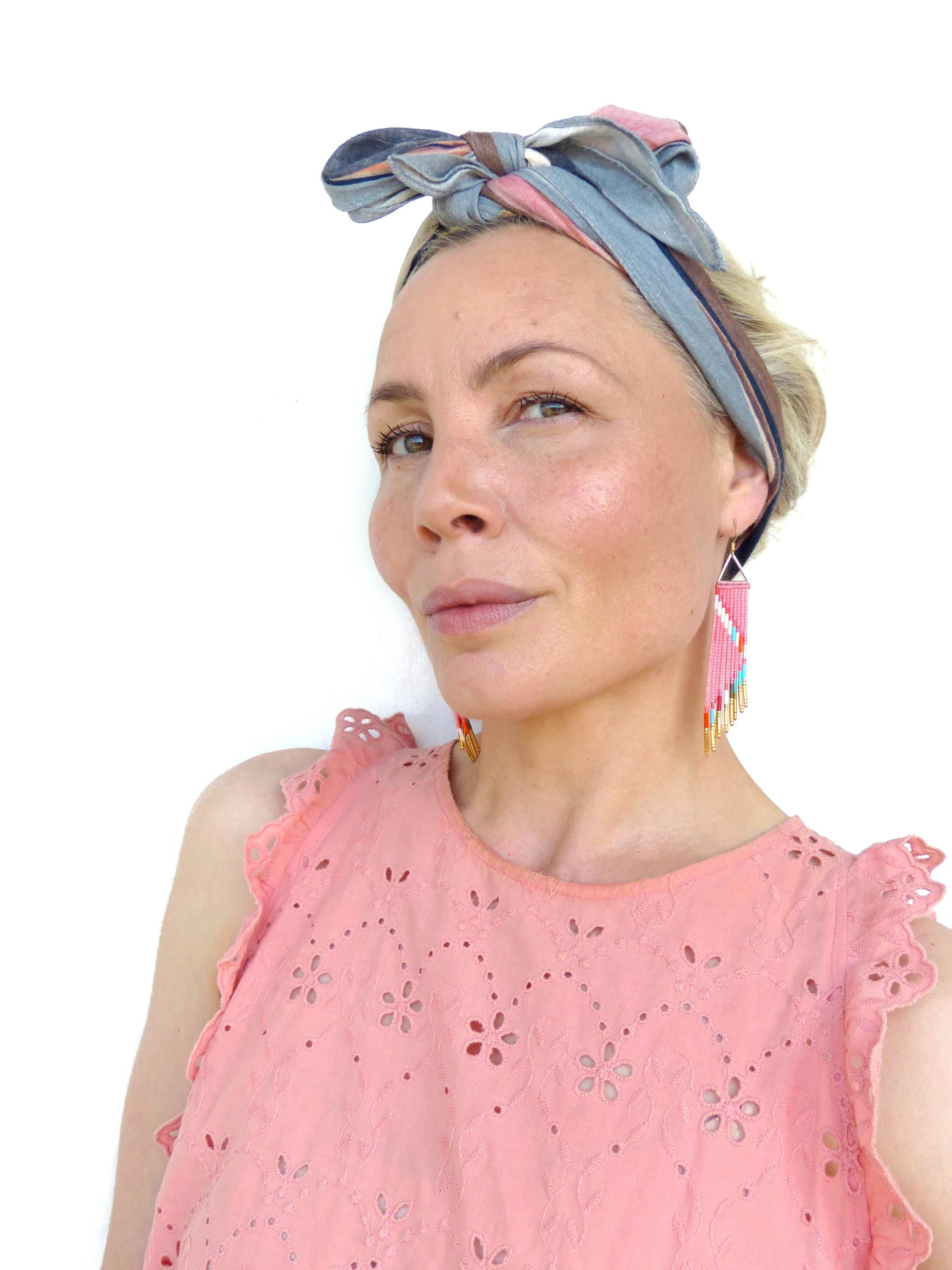 Peut inclure: Haut rose avec découpes florales et manches à volants. La personne porte un bandeau rayé et des boucles d'oreilles en perles avec des perles roses, blanches et turquoise. Les boucles d'oreilles ont un dessus triangulaire et une frange de perles colorées.