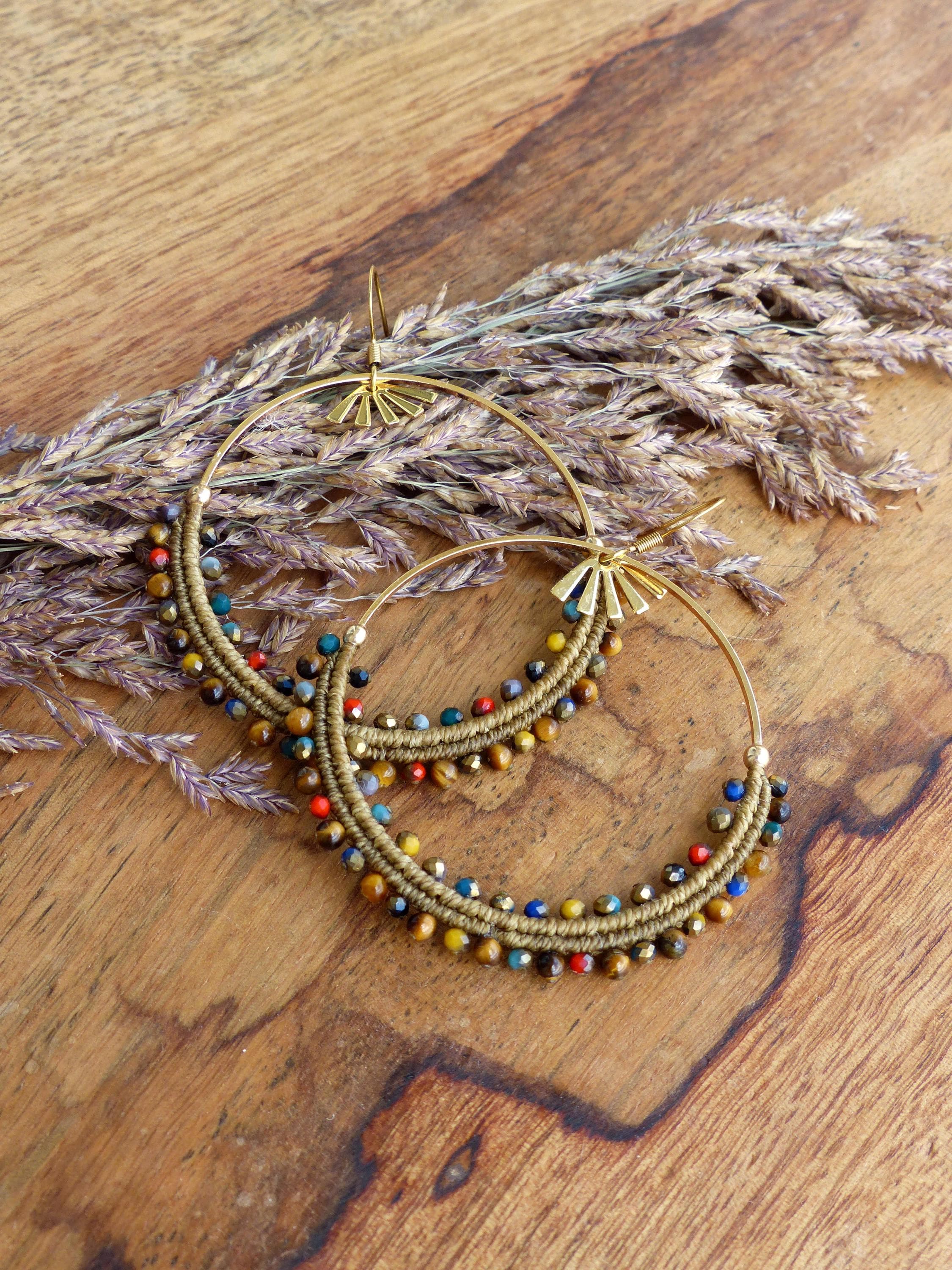 Peut inclure: Une paire de boucles d'oreilles créoles dorées avec des perles brunes, rouges, bleues et jaunes. Les boucles d'oreilles sont sur une surface en bois avec de l'herbe sèche.