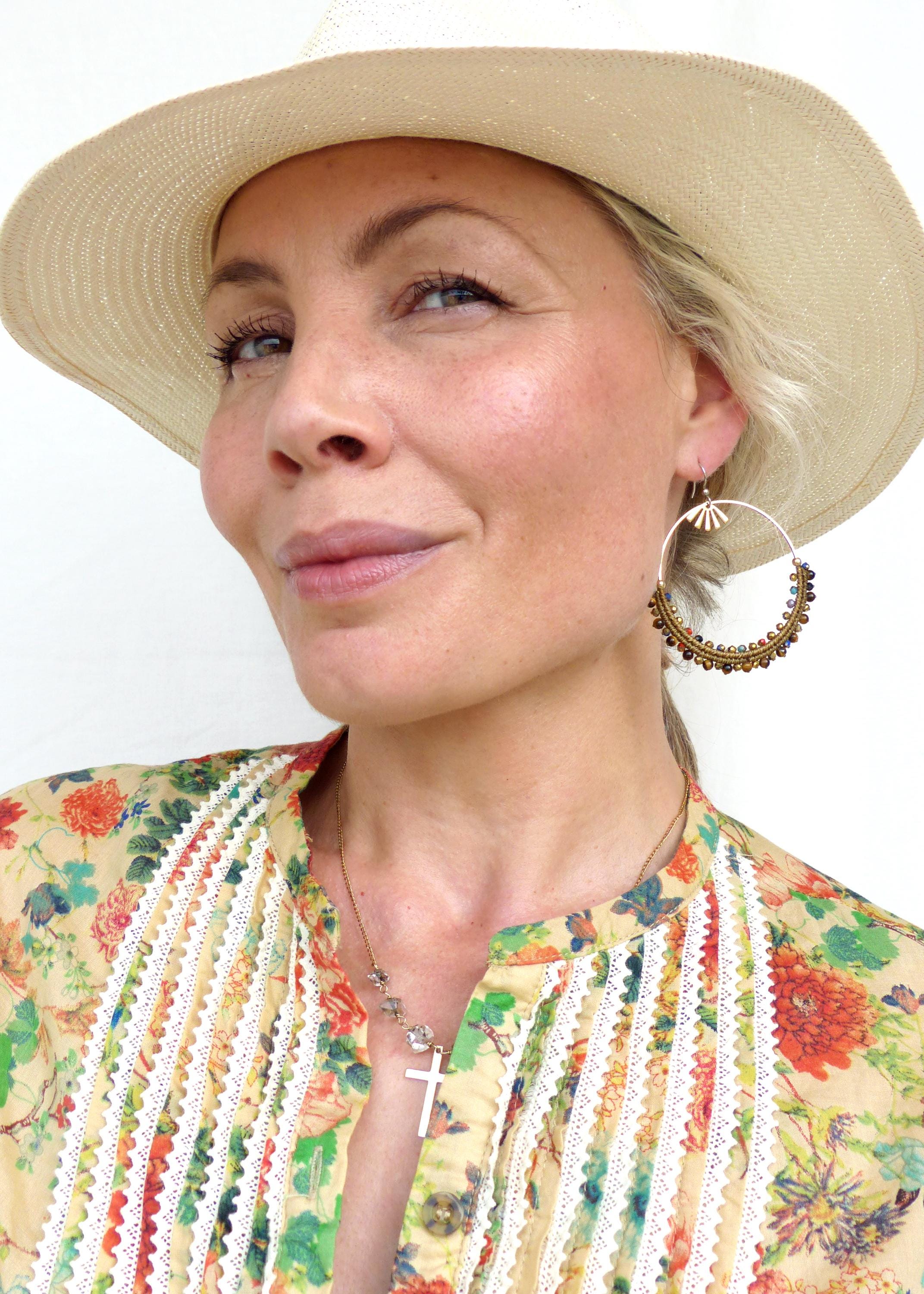 Peut inclure: Une femme portant un chapeau de paille, une blouse à imprimé floral avec des bordures en dentelle blanche et un collier en or avec un pendentif en forme de croix. Elle porte de grandes boucles d'oreilles créoles avec des perles brunes et dorées.
