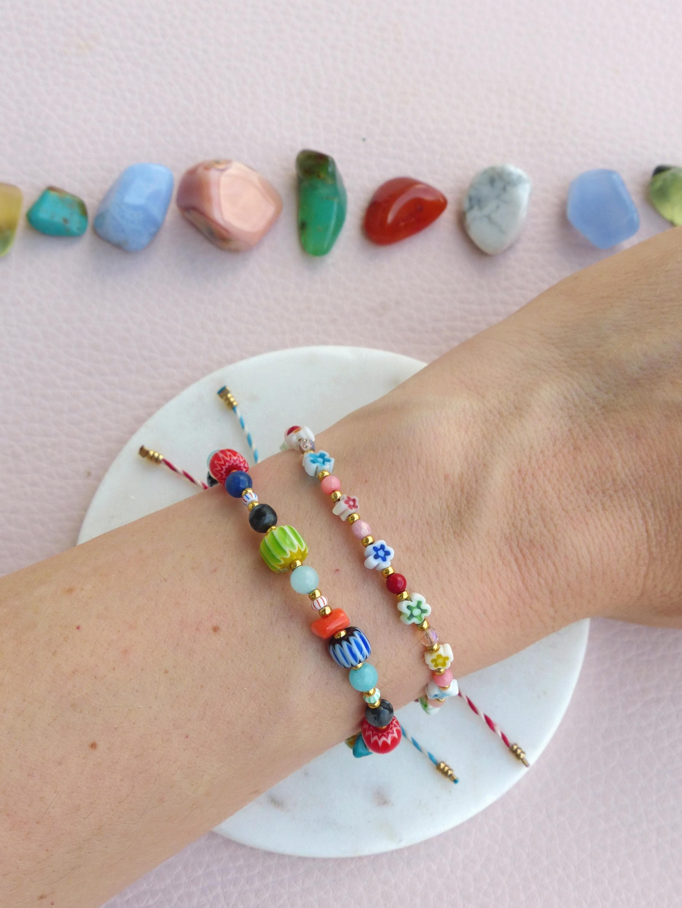 Peut inclure: Deux bracelets en perles colorées sur un poignet. Le bracelet du haut est composé de petites fleurs roses, blanches et dorées. Le bracelet du bas est composé de perles colorées variées, notamment vertes, bleues, rouges et oranges.