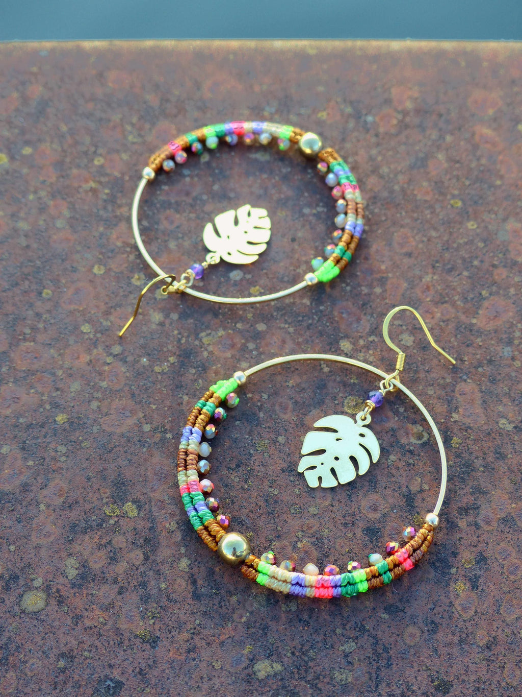 Peut inclure: Une paire de boucles d'oreilles créoles dorées avec des accents de perles colorées et un charme de feuille tropicale. Les boucles d'oreilles sont sur une surface brune.