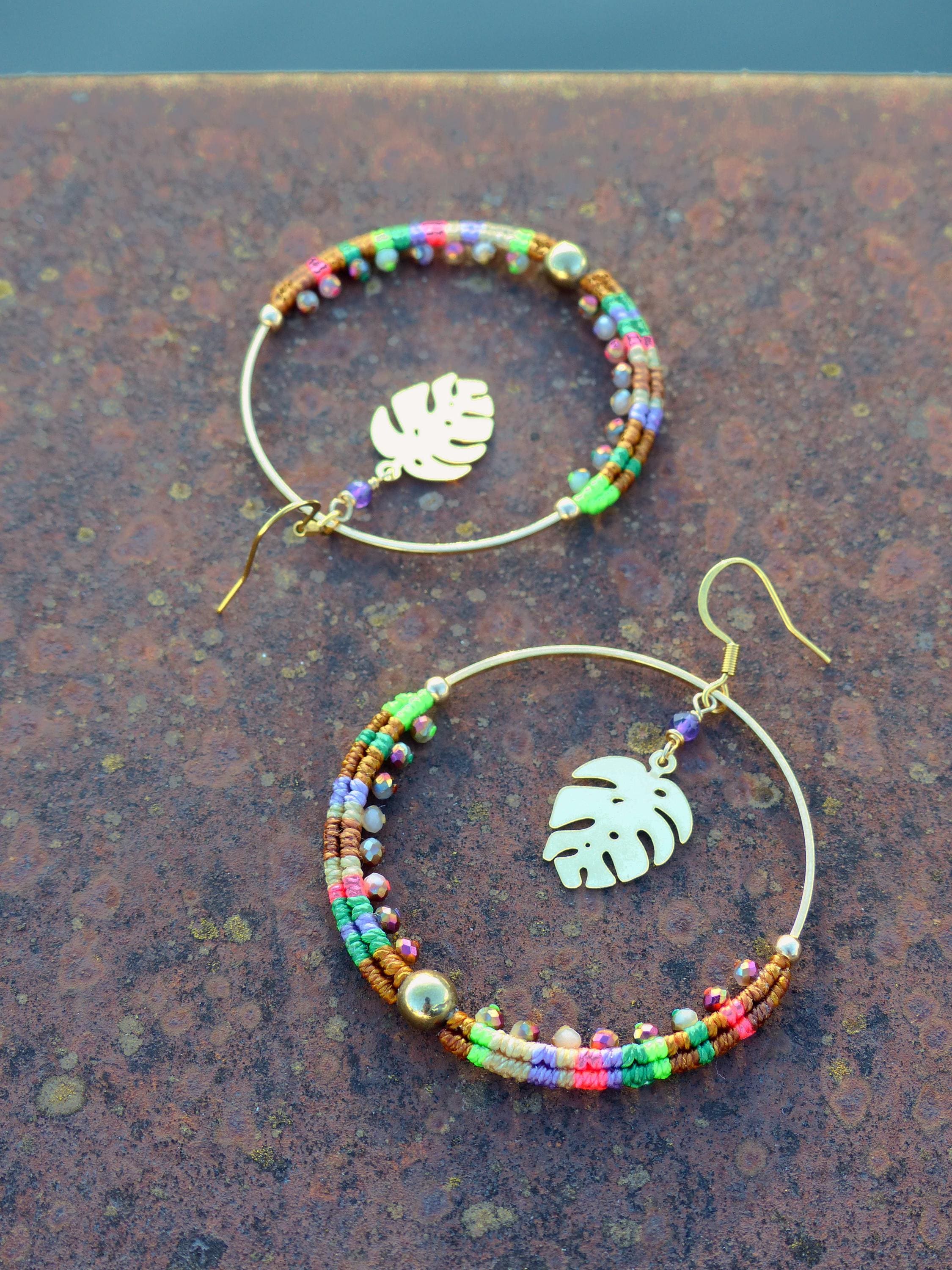 Peut inclure: Une paire de boucles d'oreilles créoles dorées avec des accents de perles colorées et un charme de feuille tropicale. Les boucles d'oreilles sont sur une surface brune.