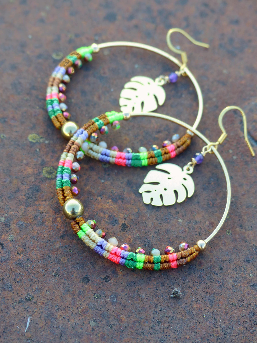 Peut inclure: Une paire de boucles d'oreilles créoles dorées avec des brins de perles colorées et un charme en forme de feuille. Les perles sont dans des tons de brun, vert, rose et violet.