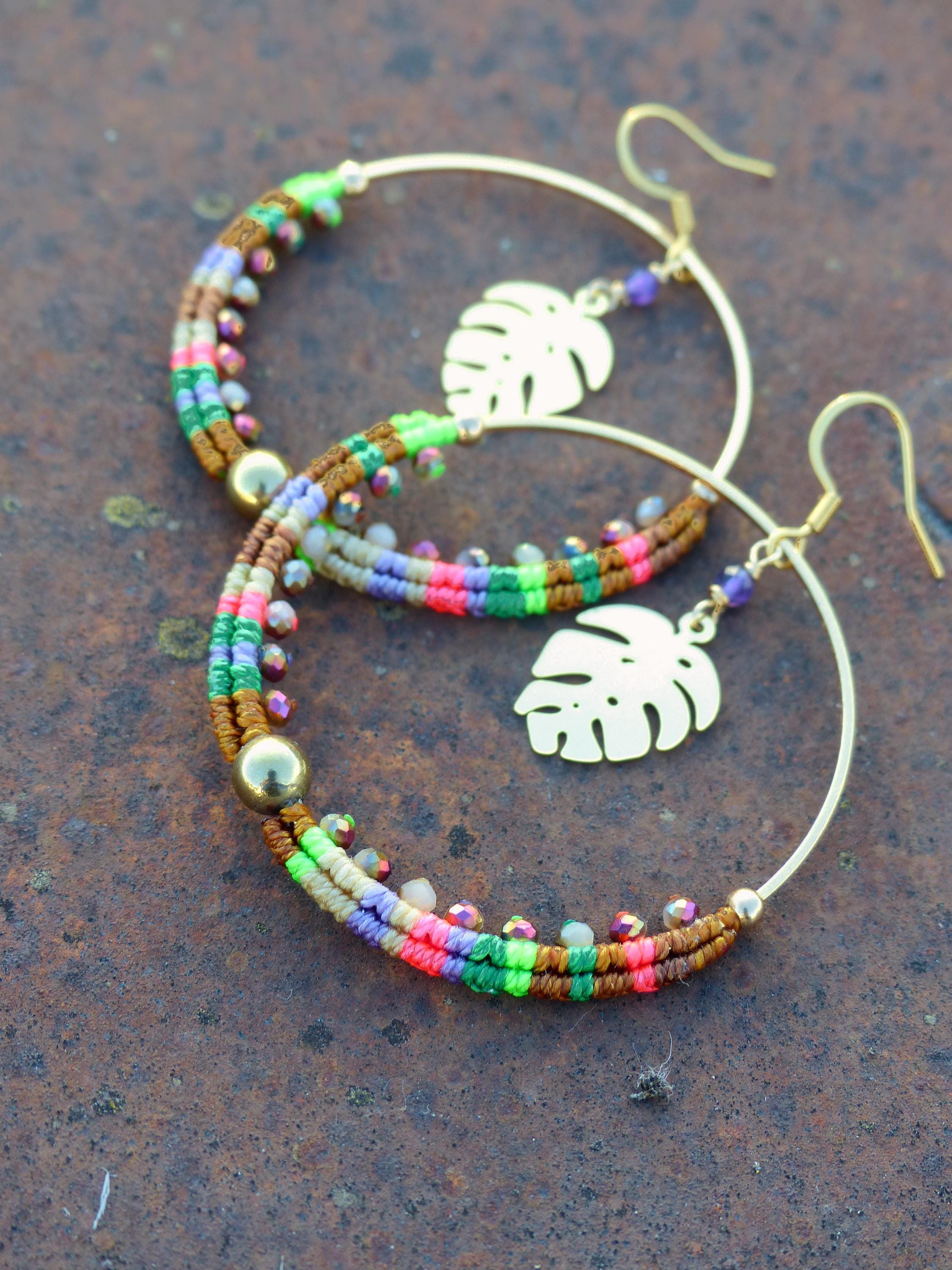 Peut inclure: Une paire de boucles d'oreilles créoles dorées avec des brins de perles colorées et un charme en forme de feuille. Les perles sont dans des tons de brun, vert, rose et violet.