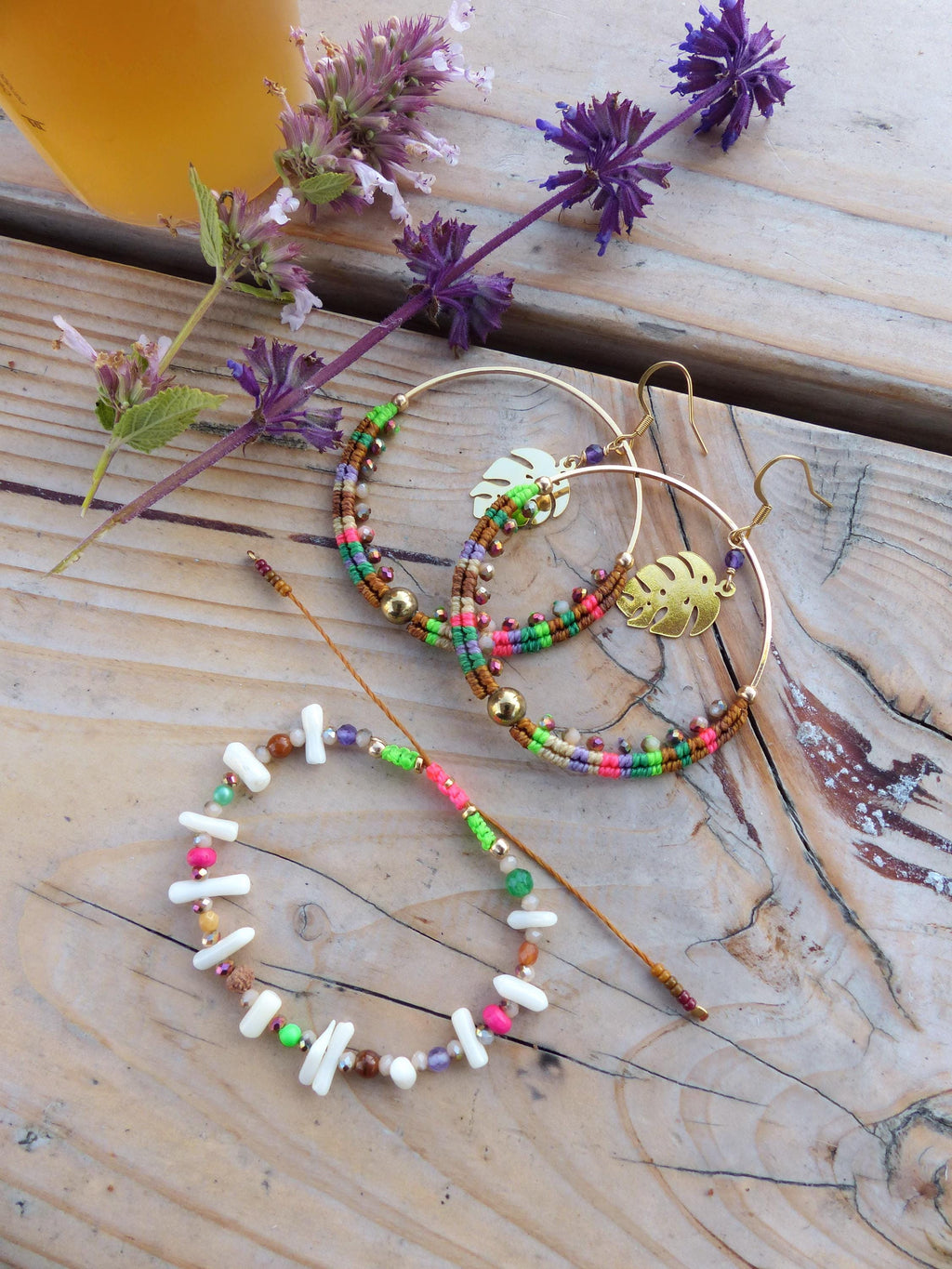 Peut inclure: Un ensemble de bijoux en perles colorées comprenant un bracelet et deux boucles d'oreilles créoles avec des accents dorés et des breloques en forme de feuilles. Les bijoux sont disposés sur une surface en bois.
