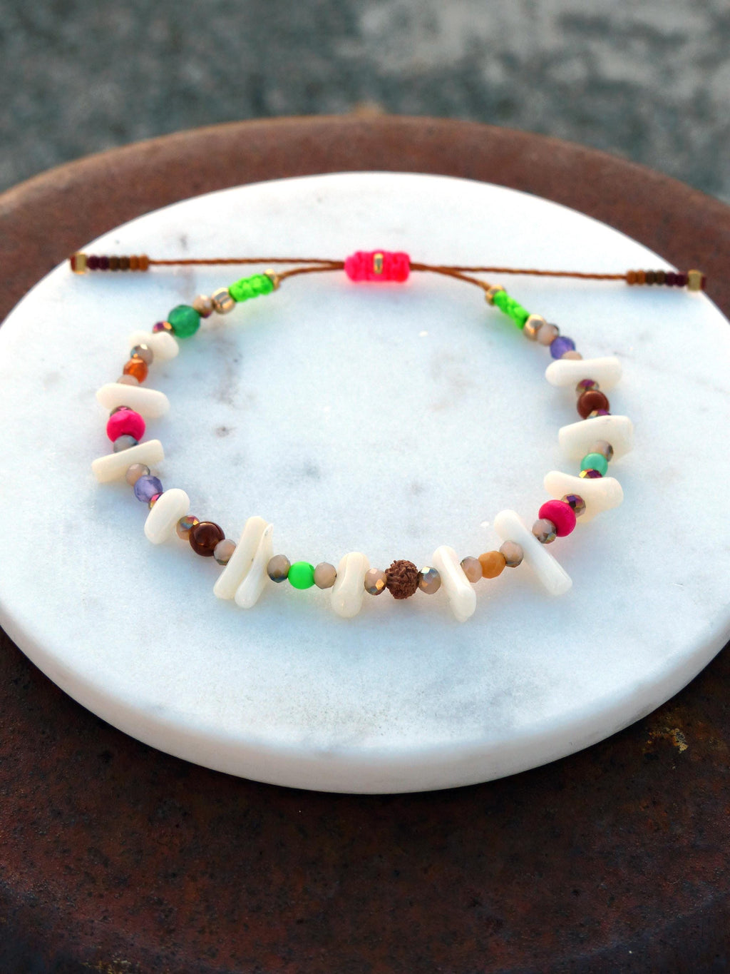 Peut inclure: Un bracelet coloré en perles avec des perles blanches, brunes, roses, vertes et violettes. Le bracelet est fait avec un cordon brun et a un nœud coulissant pour ajuster la taille.