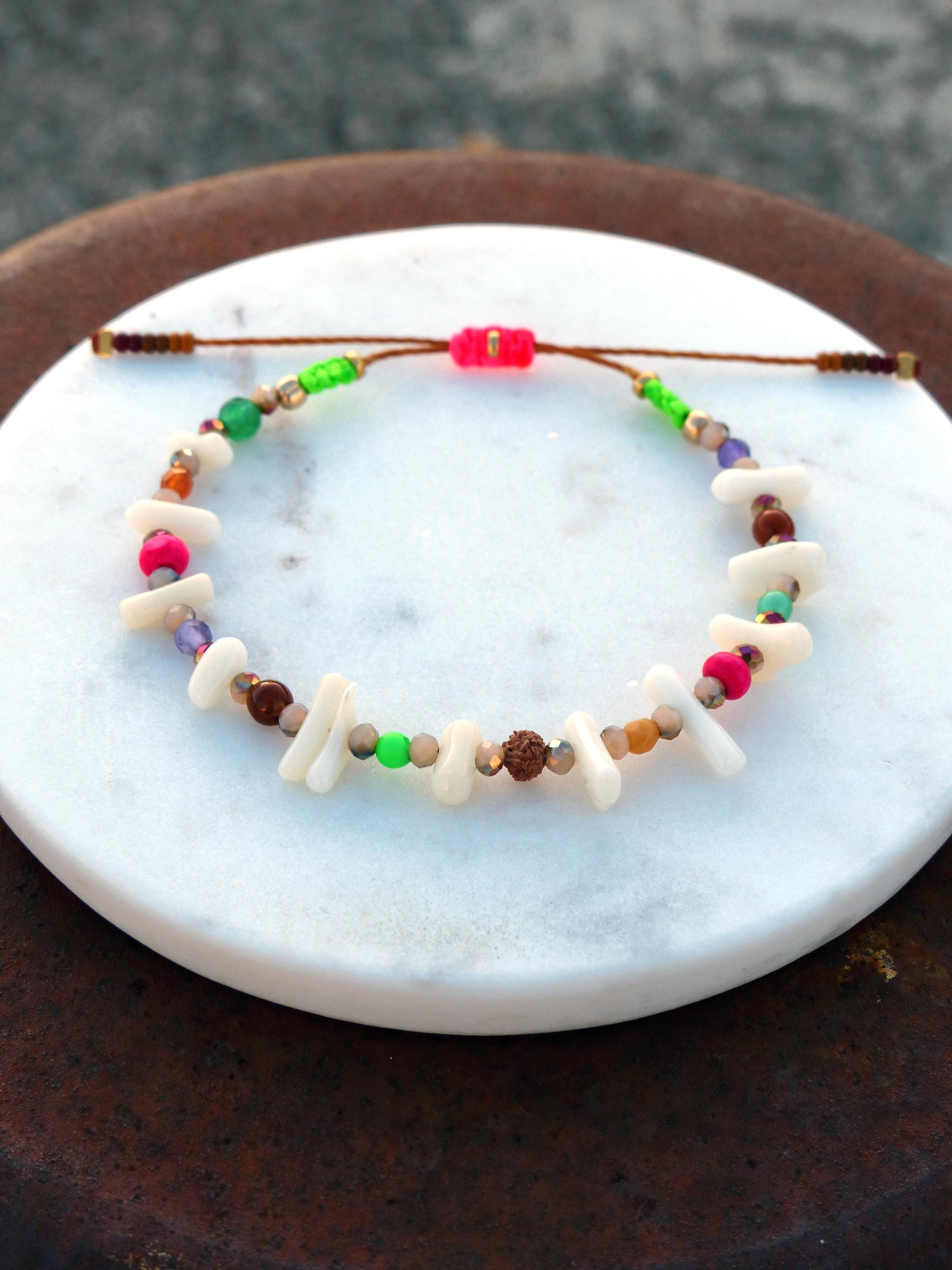 Peut inclure: Un bracelet coloré en perles avec des perles blanches, brunes, roses, vertes et violettes. Le bracelet est fait avec un cordon brun et a un nœud coulissant pour ajuster la taille.