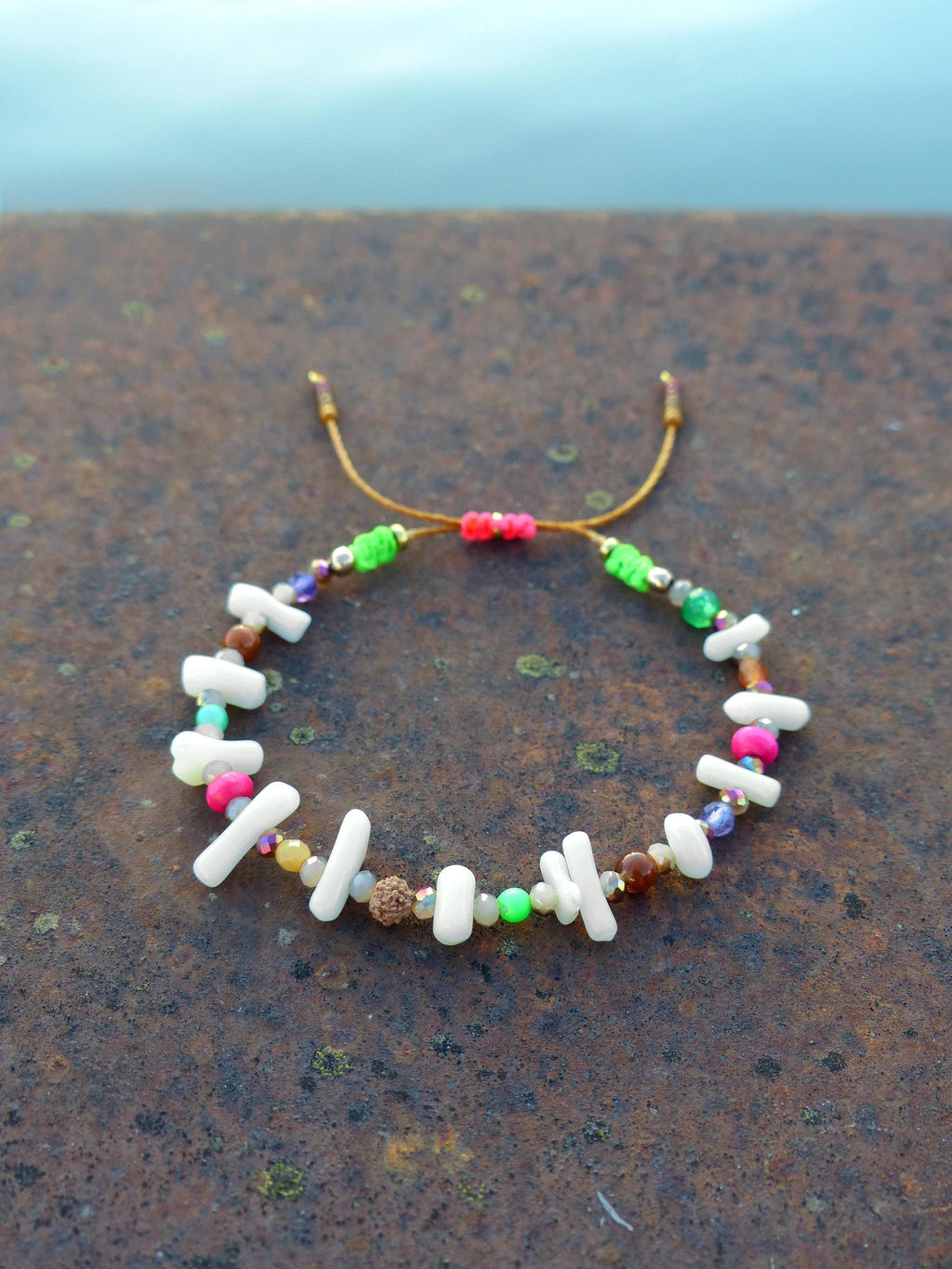 Peut inclure: Un bracelet coloré en perles avec des perles blanches, roses, vertes et brunes. Le bracelet est fait avec un cordon brun et a une fermeture à nœud.