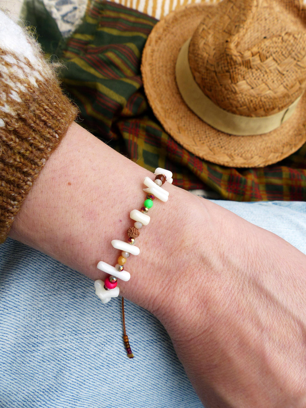Peut inclure: Un bracelet en coquillage blanc avec des perles brunes, roses, vertes et dorées. Le bracelet est attaché avec un cordon brun et est porté au poignet.