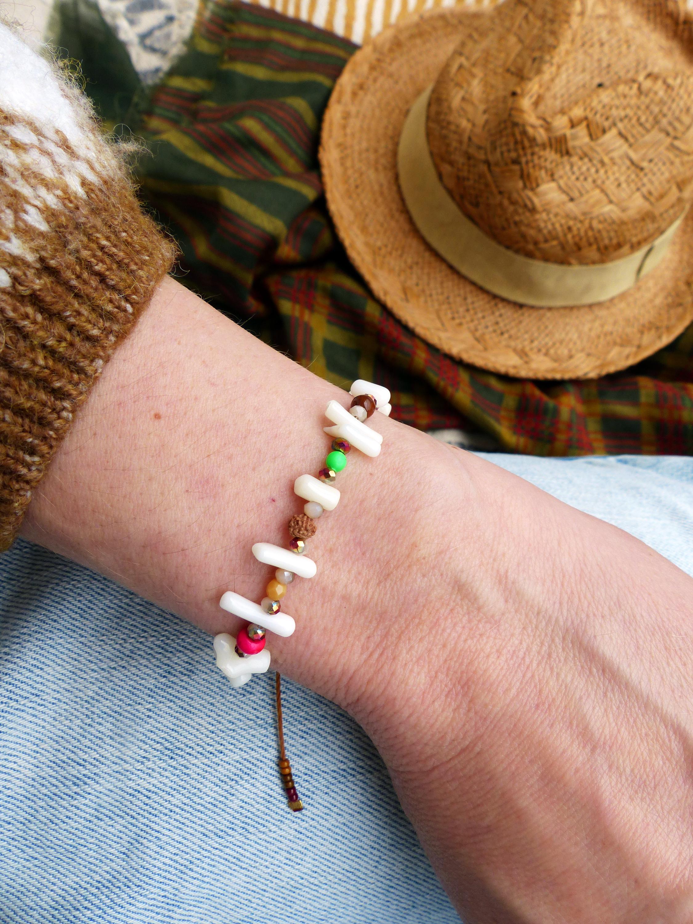 Peut inclure: Un bracelet en coquillage blanc avec des perles brunes, roses, vertes et dorées. Le bracelet est attaché avec un cordon brun et est porté au poignet.