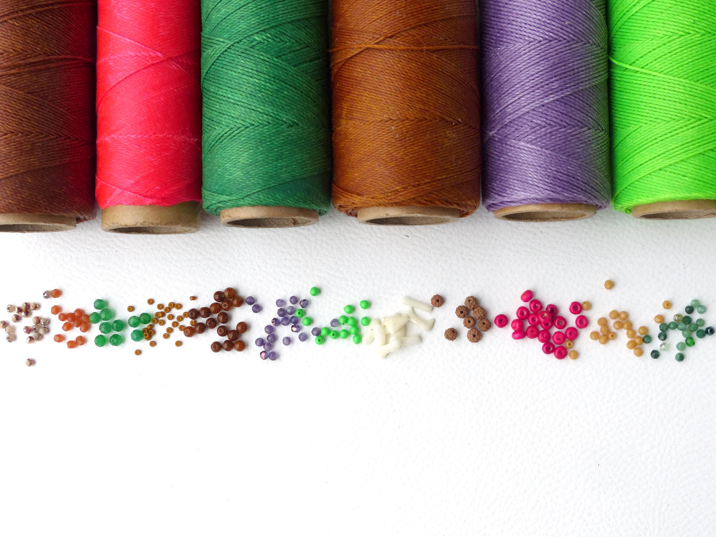 Peut inclure: Une collection de bobines de fil coloré, comprenant du brun, du rouge, du vert, du violet et du vert lime, disposées en rangée. En dessous des bobines se trouvent des rangées de petites perles colorées dans différentes nuances de brun, vert, violet, blanc, rose et vert.