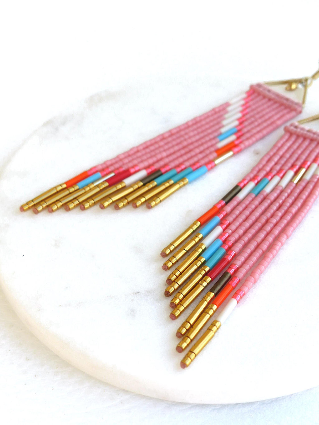 Peut inclure: Boucles d'oreilles roses en perles avec des accents dorés. Les boucles d'oreilles présentent un design à franges avec un mélange de couleurs, dont blanc, orange, rouge et bleu. Elles sont présentées sur une surface en marbre blanc.