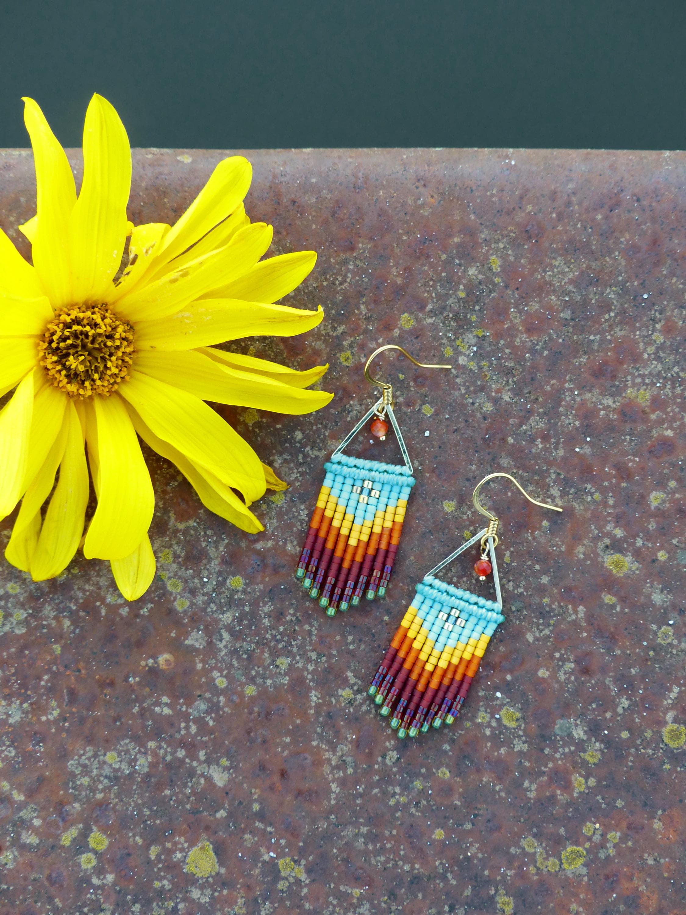 Peut inclure: Une paire de boucles d'oreilles en perles avec un schéma de couleurs turquoise, jaune, orange et rouge. Les boucles d'oreilles sont faites de petites perles et ont une forme triangulaire en haut.