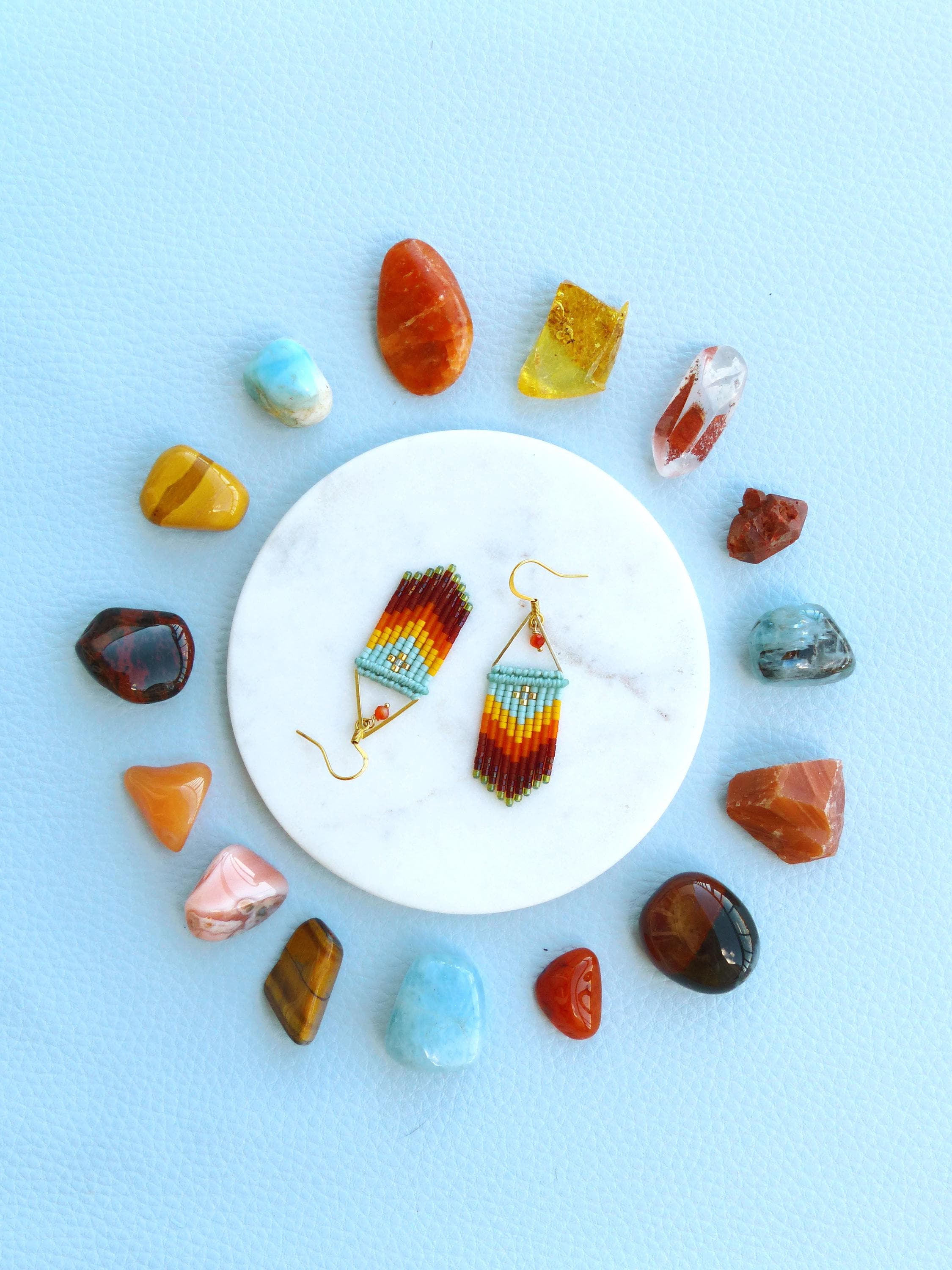 Peut inclure: Une paire de boucles d'oreilles en perles avec un motif géométrique dans des tons d'orange, rouge, jaune et bleu. Les boucles d'oreilles sont présentées sur une assiette en marbre blanc entourée de pierres de différentes couleurs.