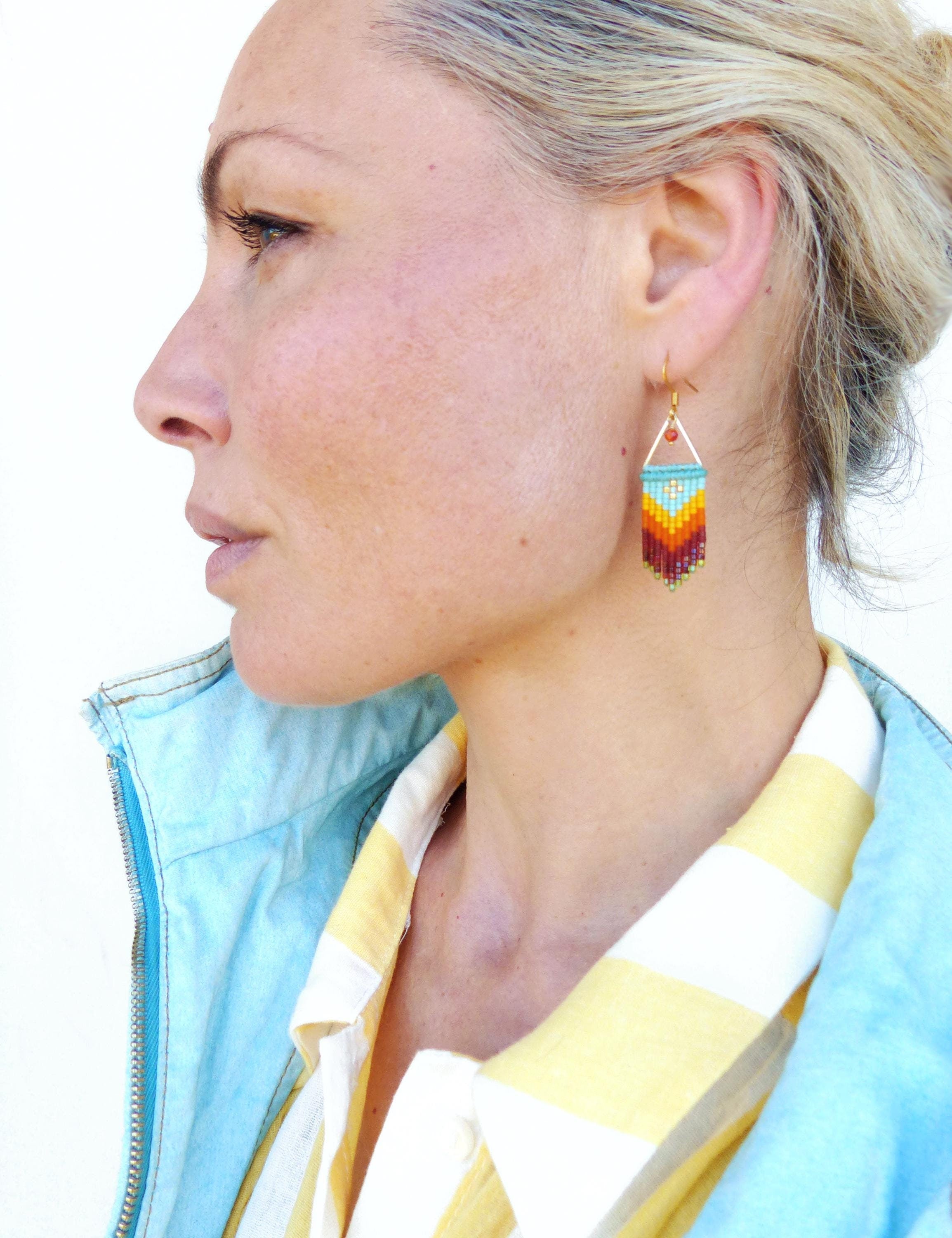 Peut inclure: Une femme portant une chemise rayée bleu et jaune avec une paire de boucles d'oreilles en perles colorées. Les boucles d'oreilles sont en forme de goutte et présentent un motif géométrique de perles rouges, orange, jaunes et bleues.