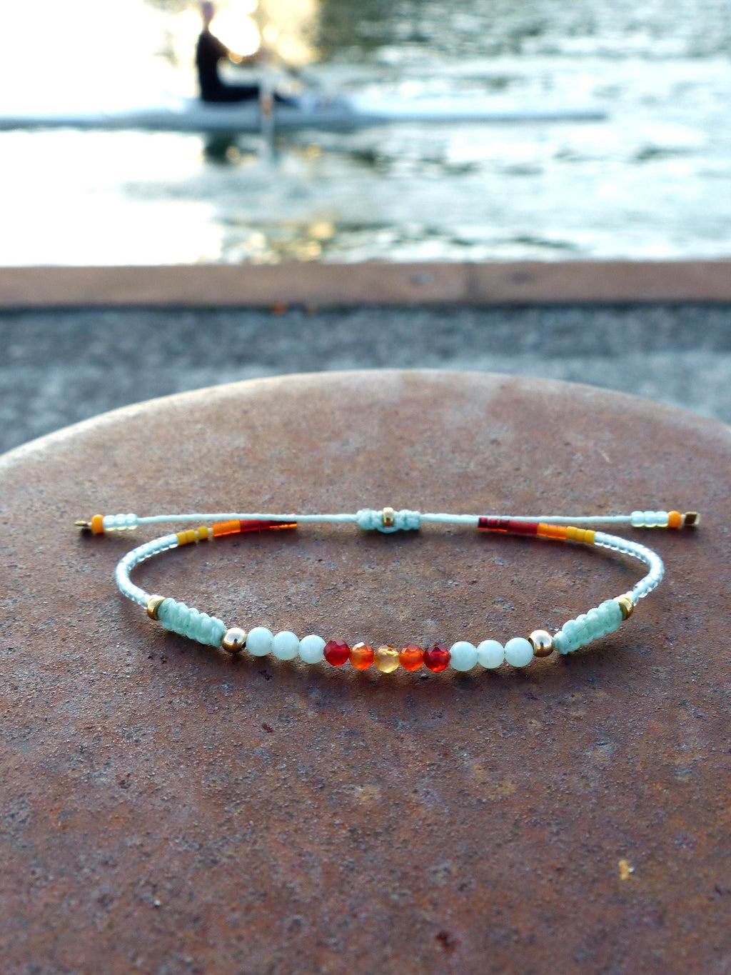 Peut inclure: Un bracelet délicat avec des perles bleu clair, orange et rouge sur un cordon blanc. Le bracelet est attaché avec un fermoir en or.