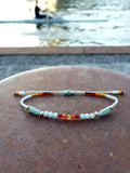 Peut inclure: Un bracelet délicat avec des perles bleu clair, orange et rouge sur un cordon blanc. Le bracelet est attaché avec un fermoir en or.