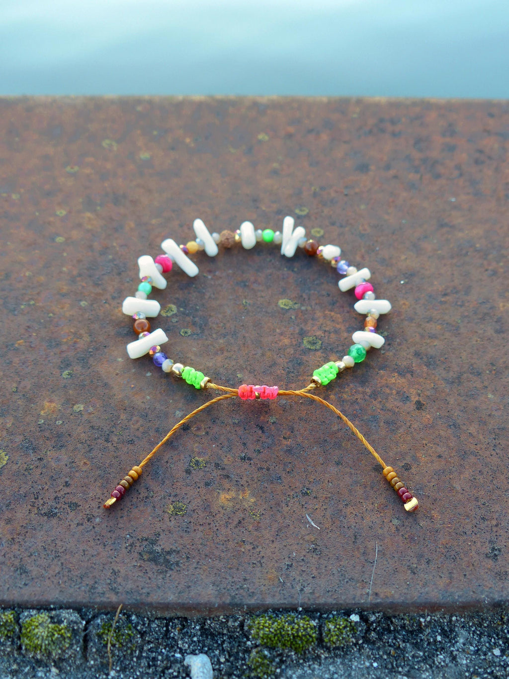 Peut inclure: Un bracelet coloré composé de corail blanc, de perles brunes, vertes, roses et violettes. Le bracelet est noué avec un cordon brun.