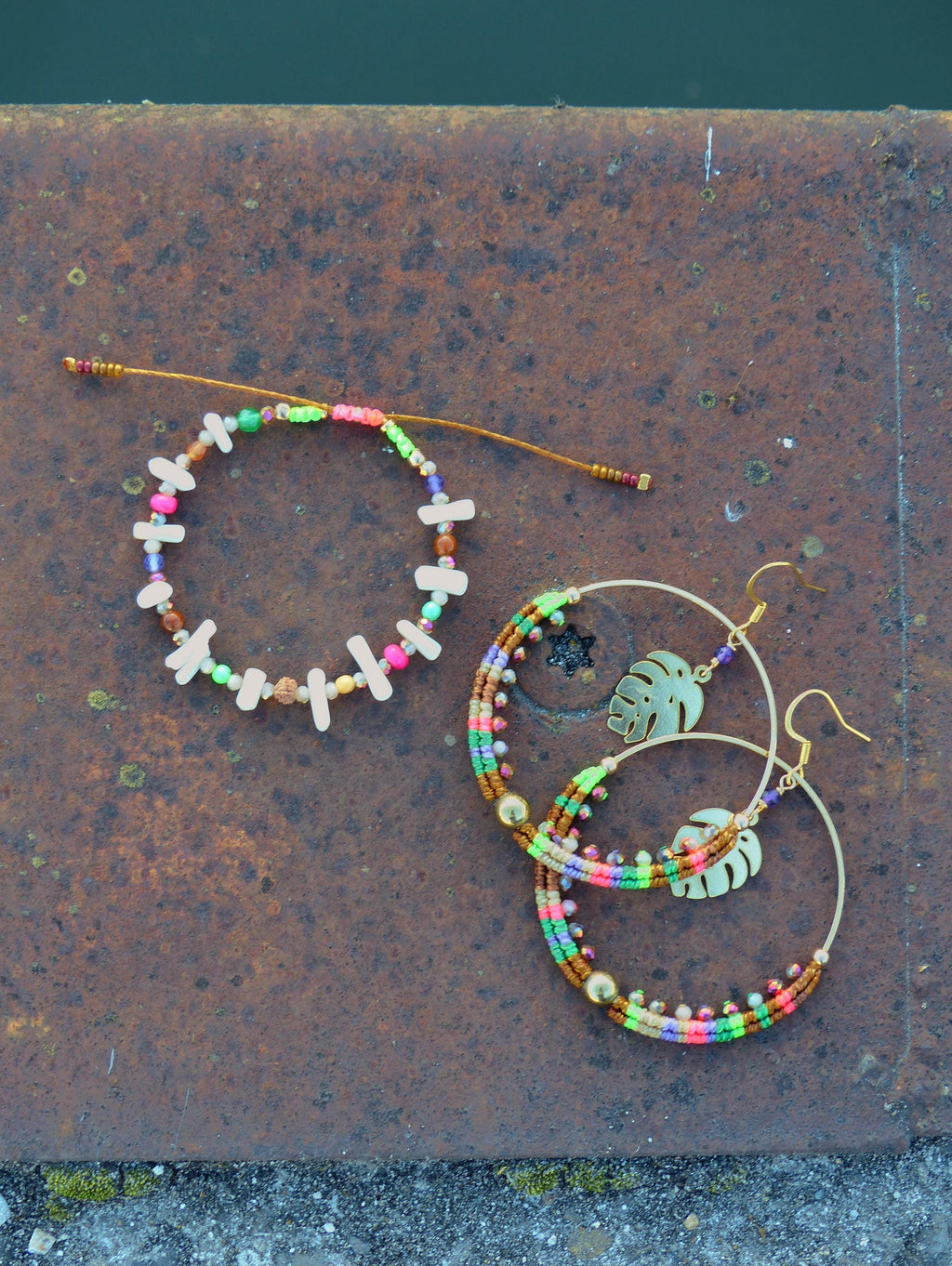 Peut inclure: Deux paires de boucles d'oreilles en perles colorées avec des cercles dorés et un charme de feuille tropicale. Les boucles d'oreilles sont sur une surface brun rouille.