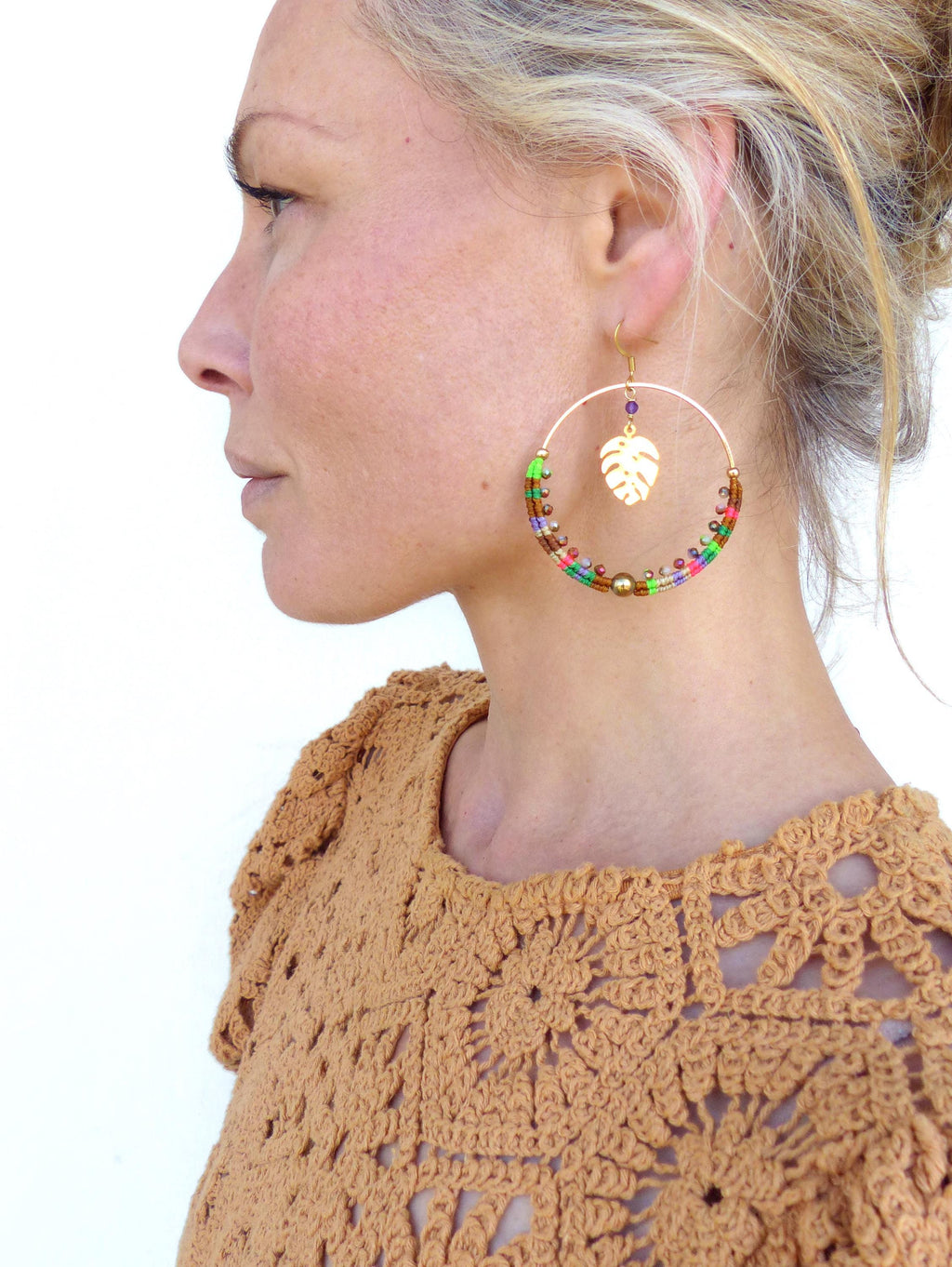 Peut inclure: Une paire de grandes boucles d'oreilles créoles avec un cadre en métal doré et des perles colorées. Un charme en feuille d'or pend au bas du cerceau.