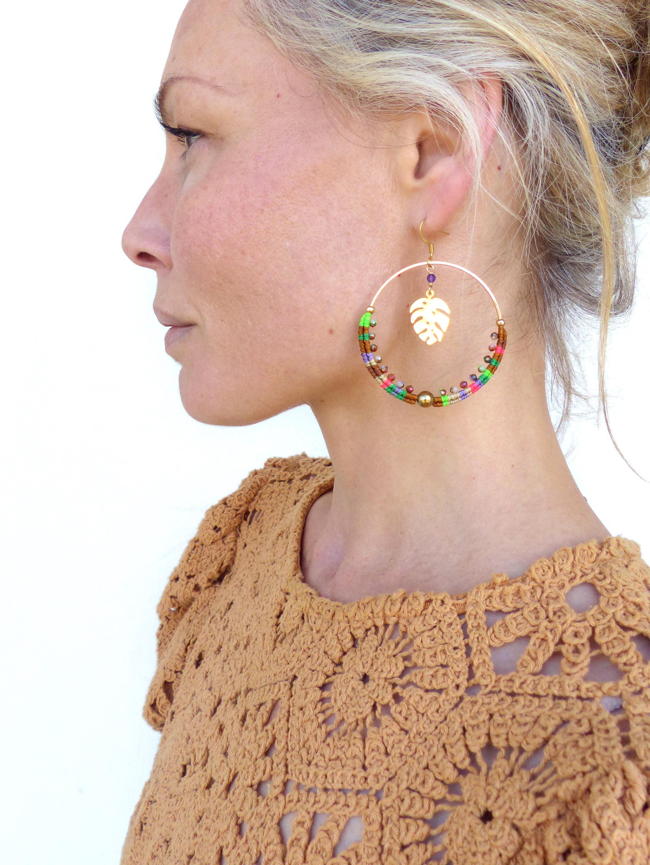 Peut inclure: Une paire de grandes boucles d'oreilles créoles avec un cadre en métal doré et des perles colorées. Un charme en feuille d'or pend au bas du cerceau.
