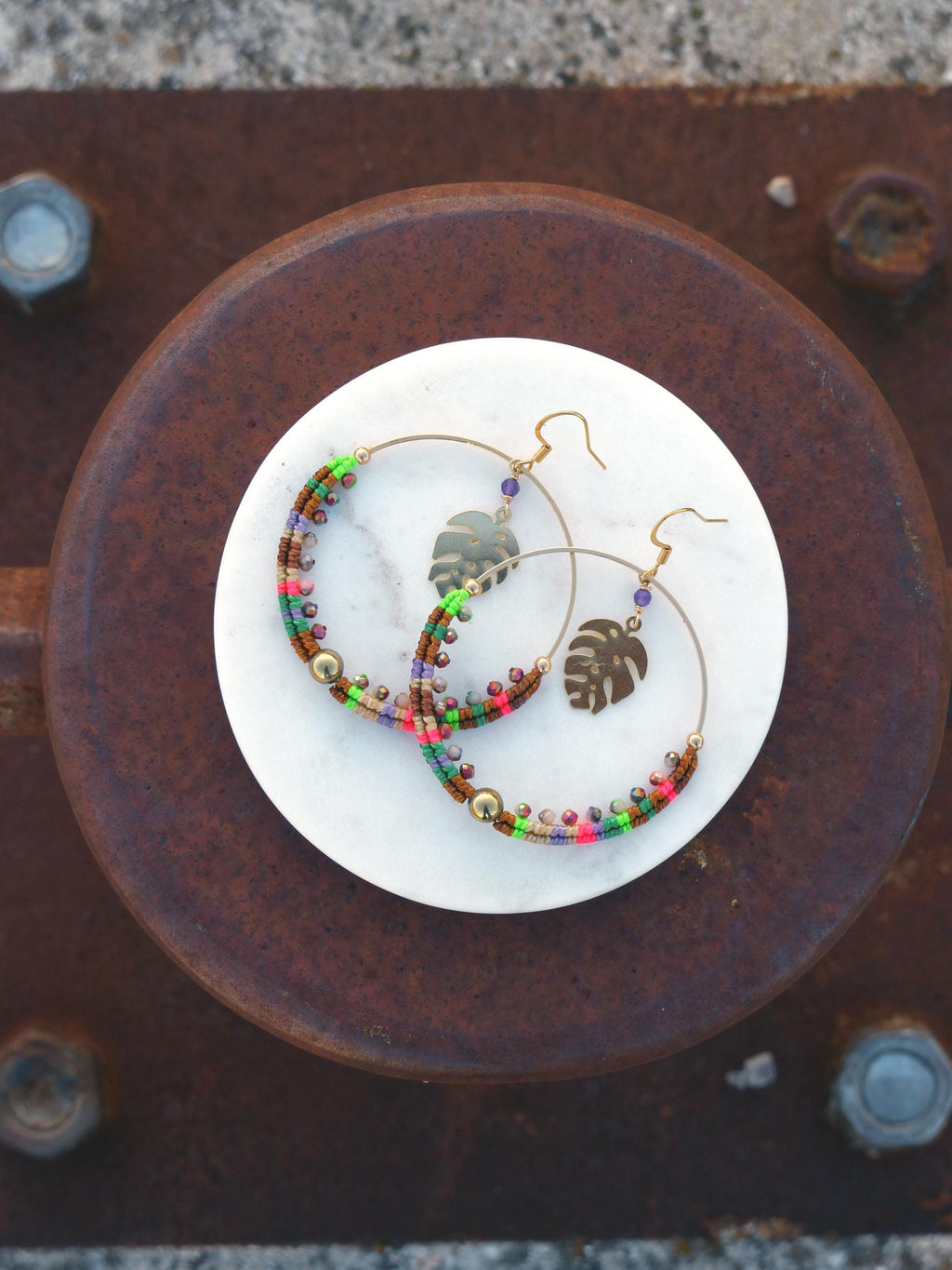 Peut inclure: Une paire de boucles d'oreilles créoles dorées avec des perles colorées et un charme en forme de feuille. Les boucles d'oreilles sont sur une surface blanche.