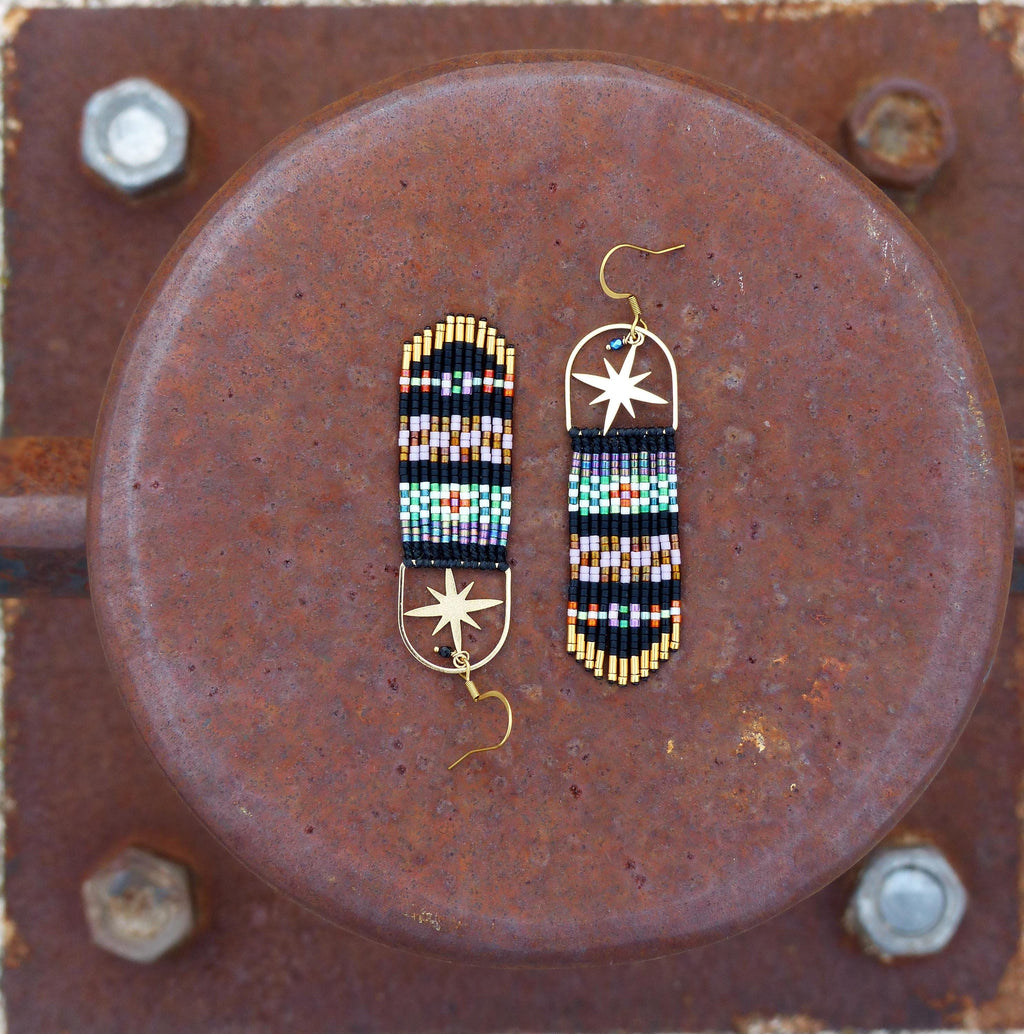 Peut inclure: Une paire de longues boucles d'oreilles en perles avec un charme d'étoile dorée. Les boucles d'oreilles sont noires avec des motifs géométriques colorés et des accents dorés.
