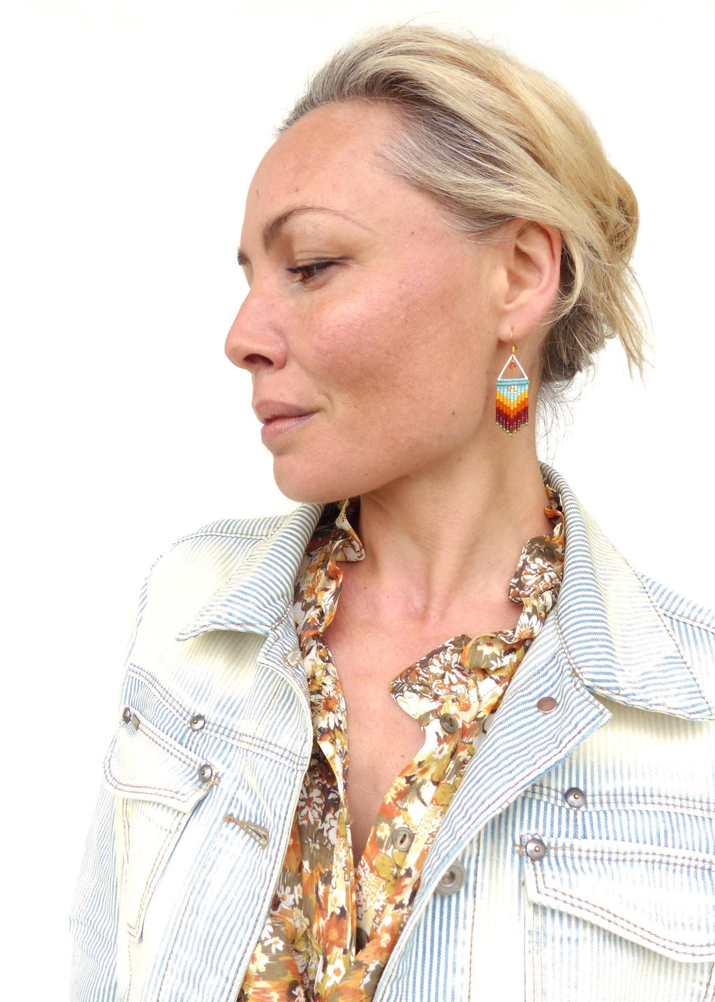 Peut inclure: Une femme portant une veste en jean rayée sur une blouse à fleurs avec une paire de boucles d'oreilles en perles colorées. Les boucles d'oreilles sont de forme triangulaire et présentent un dégradé de perles orange, jaune, bleu et vert.