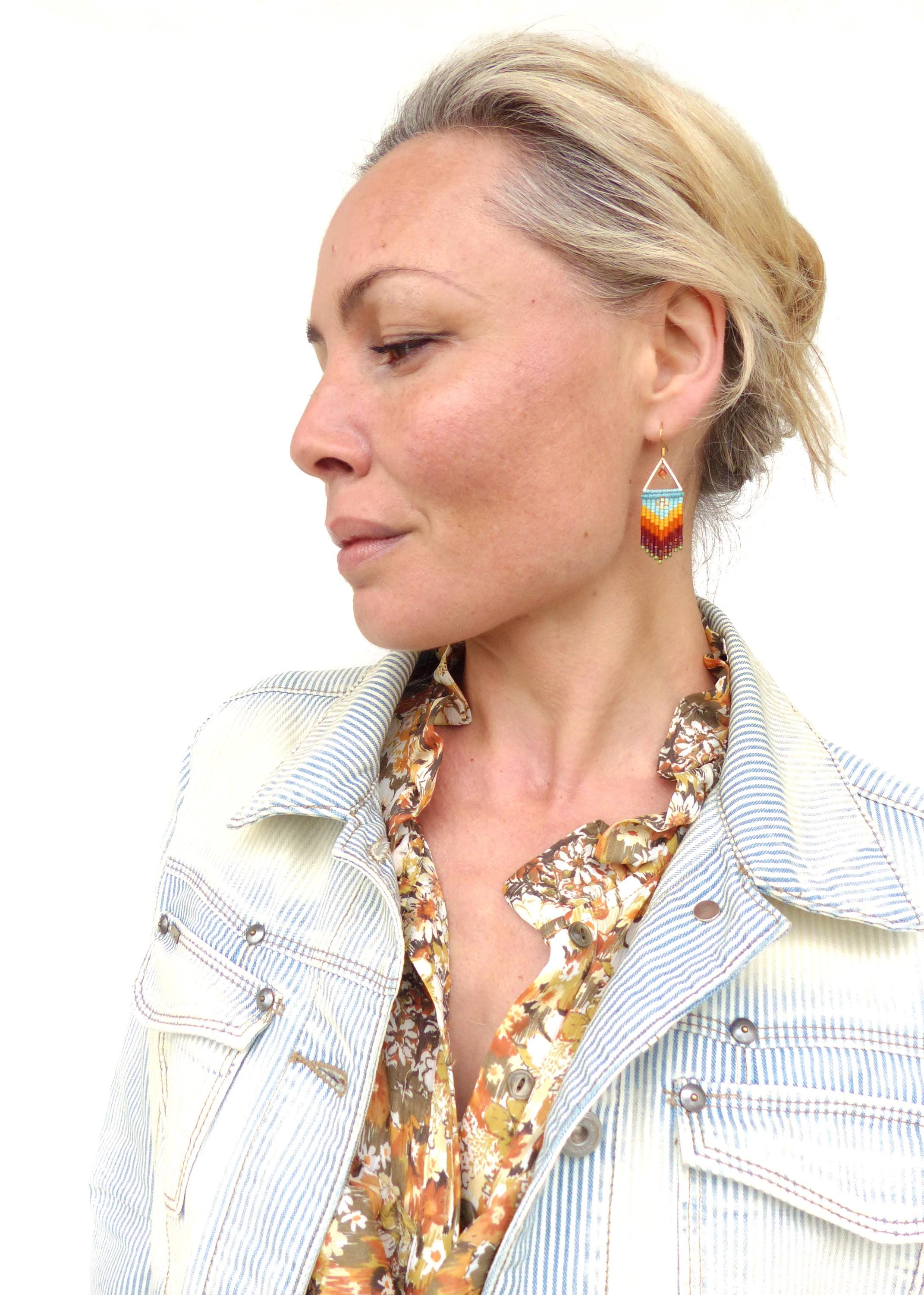 Peut inclure: Une femme portant une veste en jean rayée sur une blouse à fleurs avec une paire de boucles d'oreilles en perles colorées. Les boucles d'oreilles sont de forme triangulaire et présentent un dégradé de perles orange, jaune, bleu et vert.