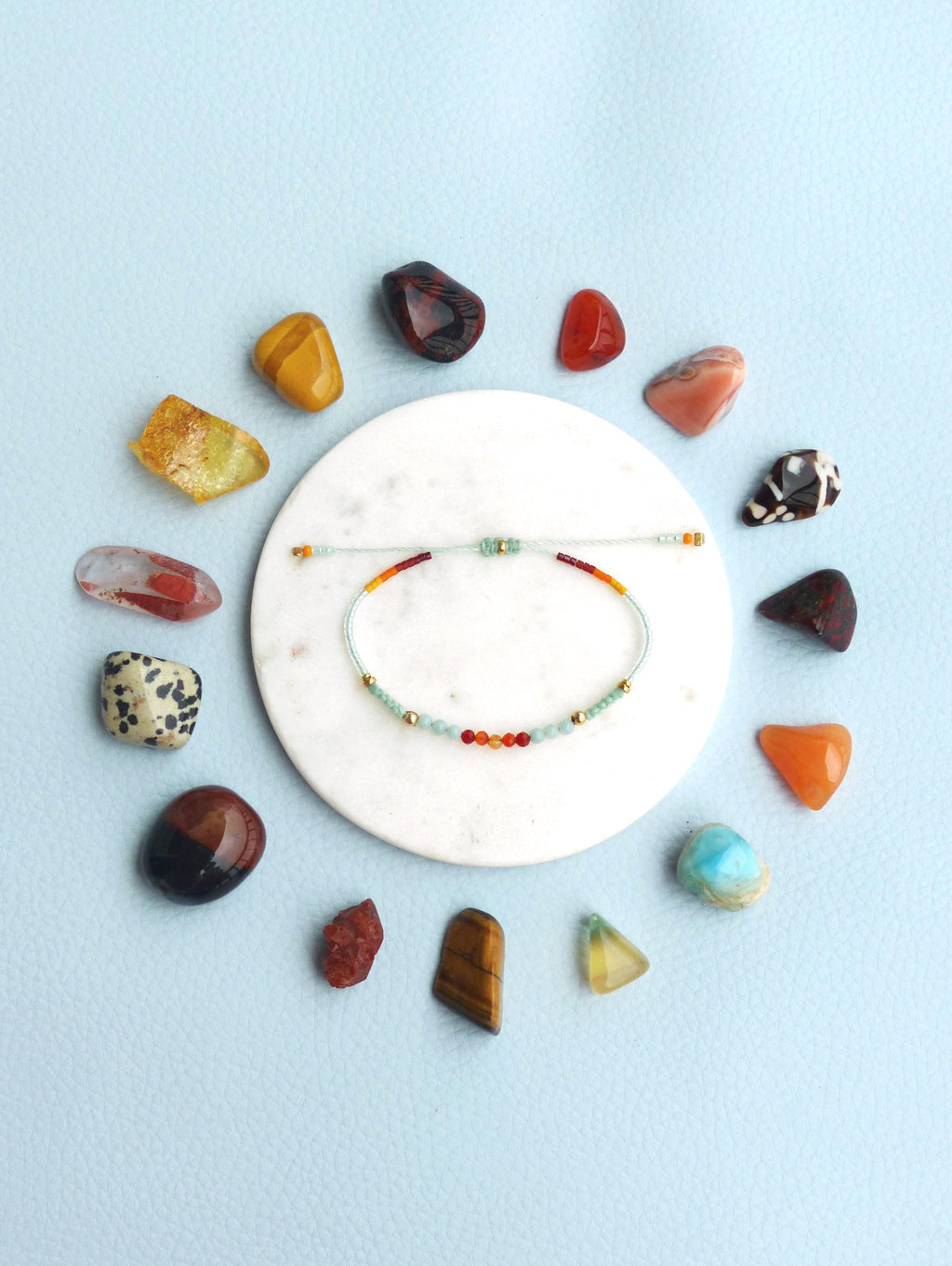 Peut inclure: Un bracelet en perles colorées avec un motif bleu clair, orange, rouge et jaune. Le bracelet est sur une assiette blanche entourée de différentes pierres.