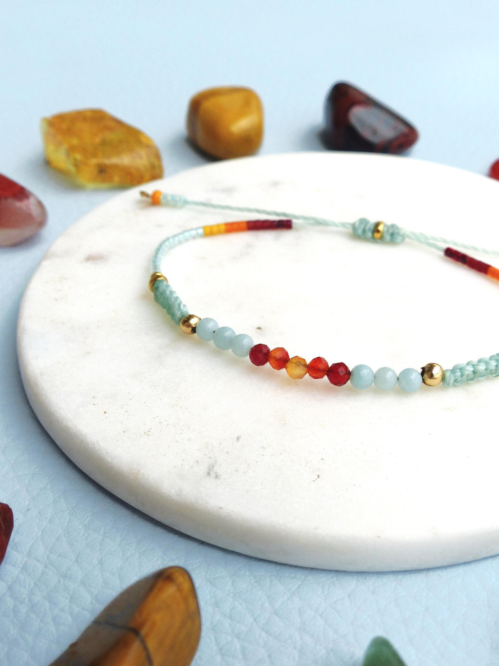 Peut inclure: Un bracelet délicat avec un arc-en-ciel de petites perles, dont des orange, rouge et bleu clair, sur un cordon blanc. Le bracelet est sur une surface en marbre blanc.
